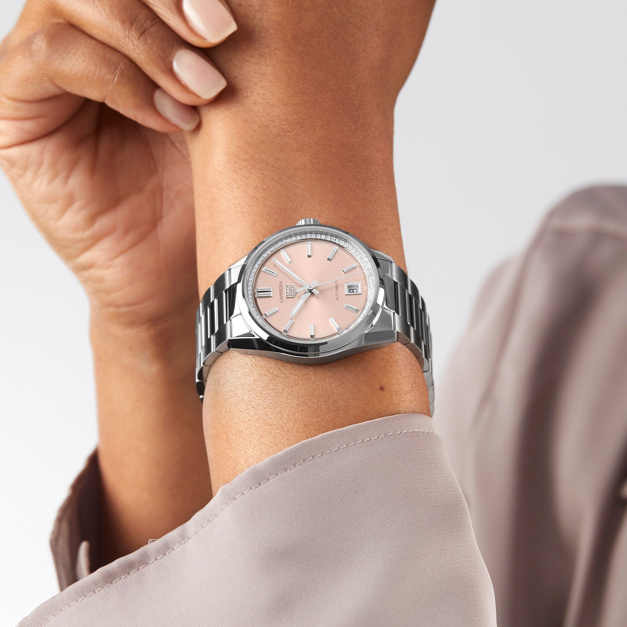 Wearing a TAG Heuer Carrera Date wristwatch with a light pink dial and date display, someone in a light grey long-sleeve garment holds their own wrist.