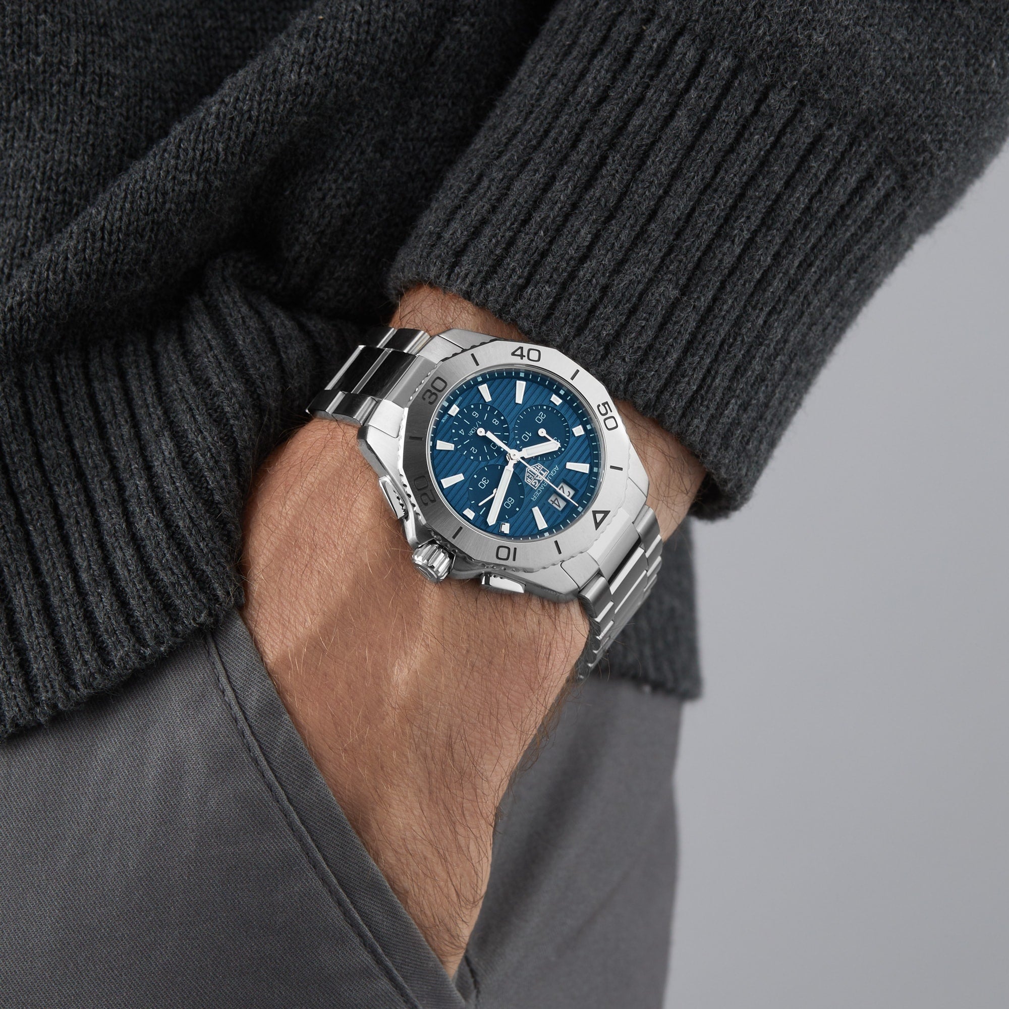 A person wears a TAG Heuer Aquaracer 200 Professional Solargraph, featuring a silver bracelet and blue face, with hand in pocket.