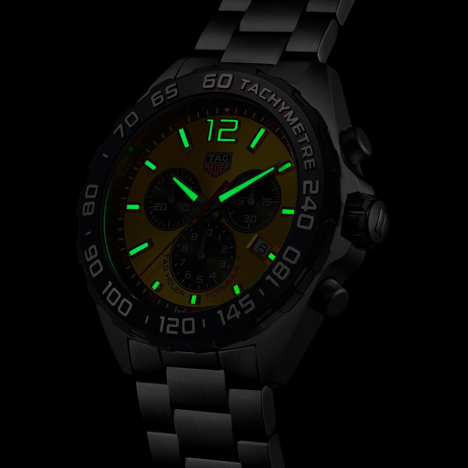 The TAG Heuer Formula 1 Chronograph, with an illuminated dial and hands, tachymeter bezel, and metal link band, shines in low light—capturing the motor racing spirit.