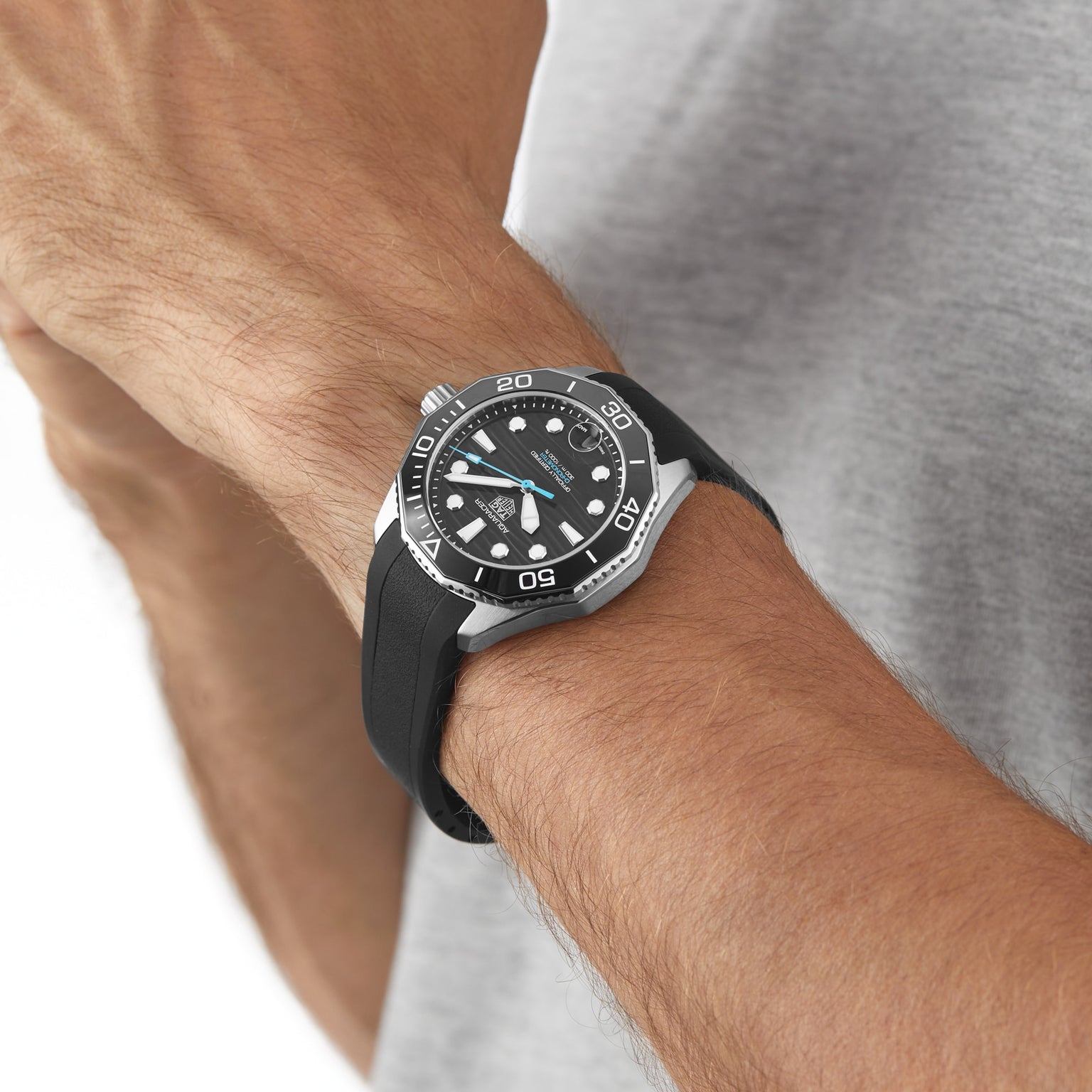 A person wearing a TAG Heuer Aquaracer Professional 300 Date with a rubber strap on their wrist, dressed in a grey shirt.