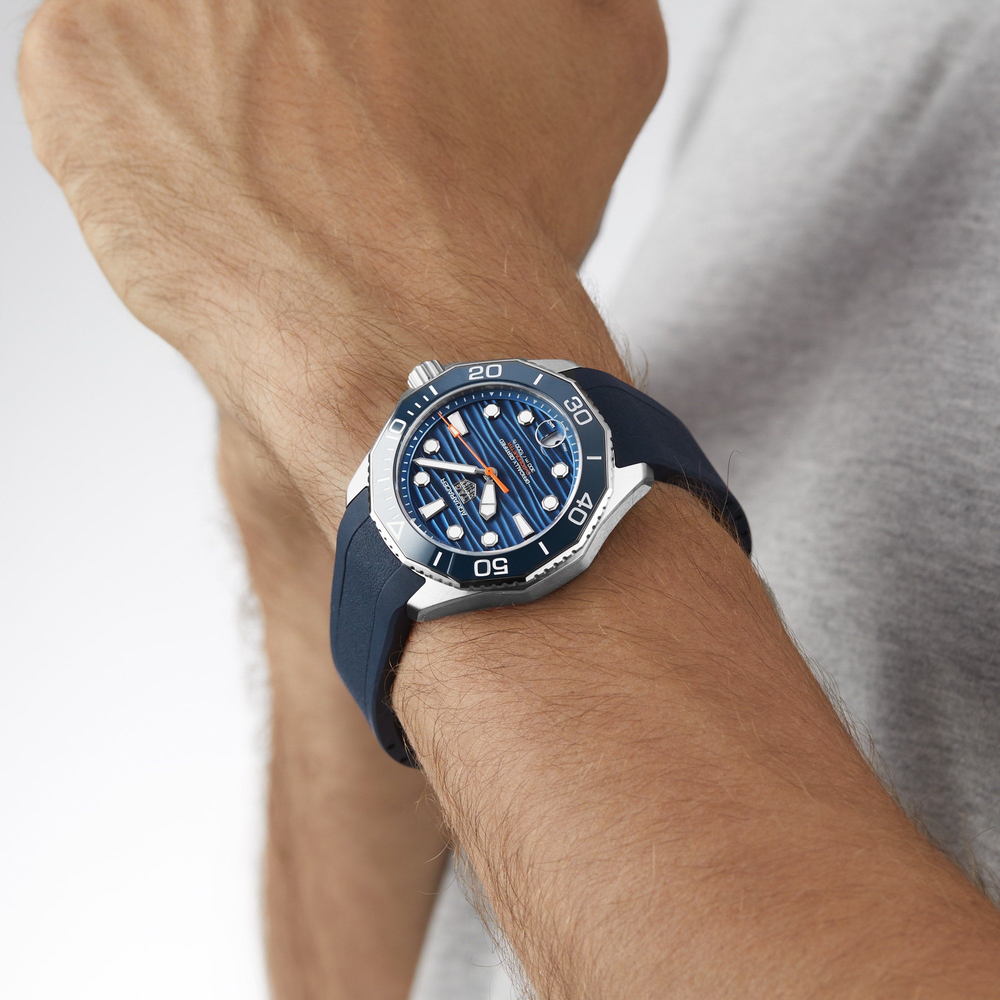 A person wears a TAG Heuer Aquaracer Professional 300 Date with a blue strap and striped dial on their left wrist against a plain background—a luxury chronograph inspired by deep-sea exploration.