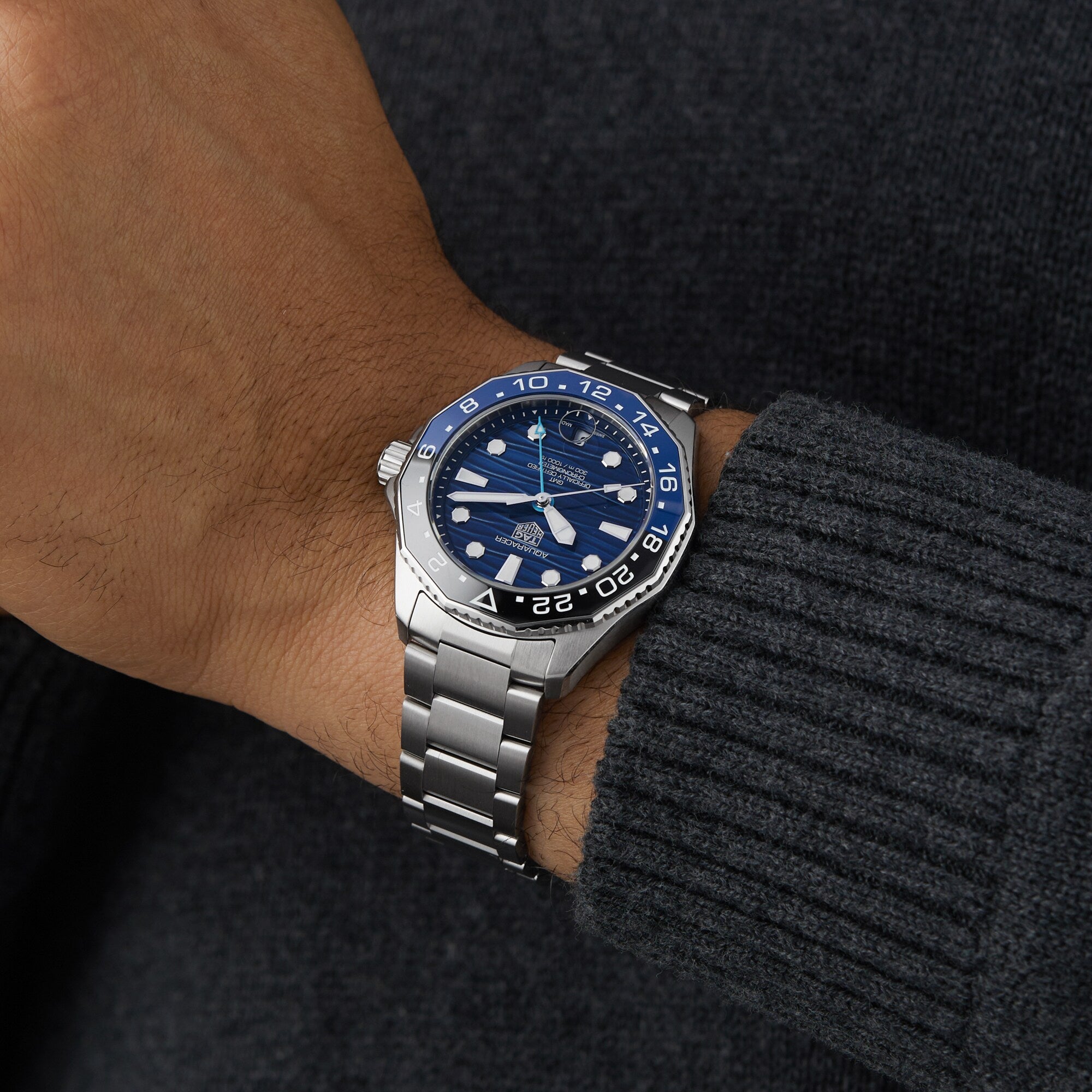 A person wearing a TAG Heuer Aquaracer Professional 300 GMT silver wristwatch with a blue and black face, dressed in a dark gray sweater.