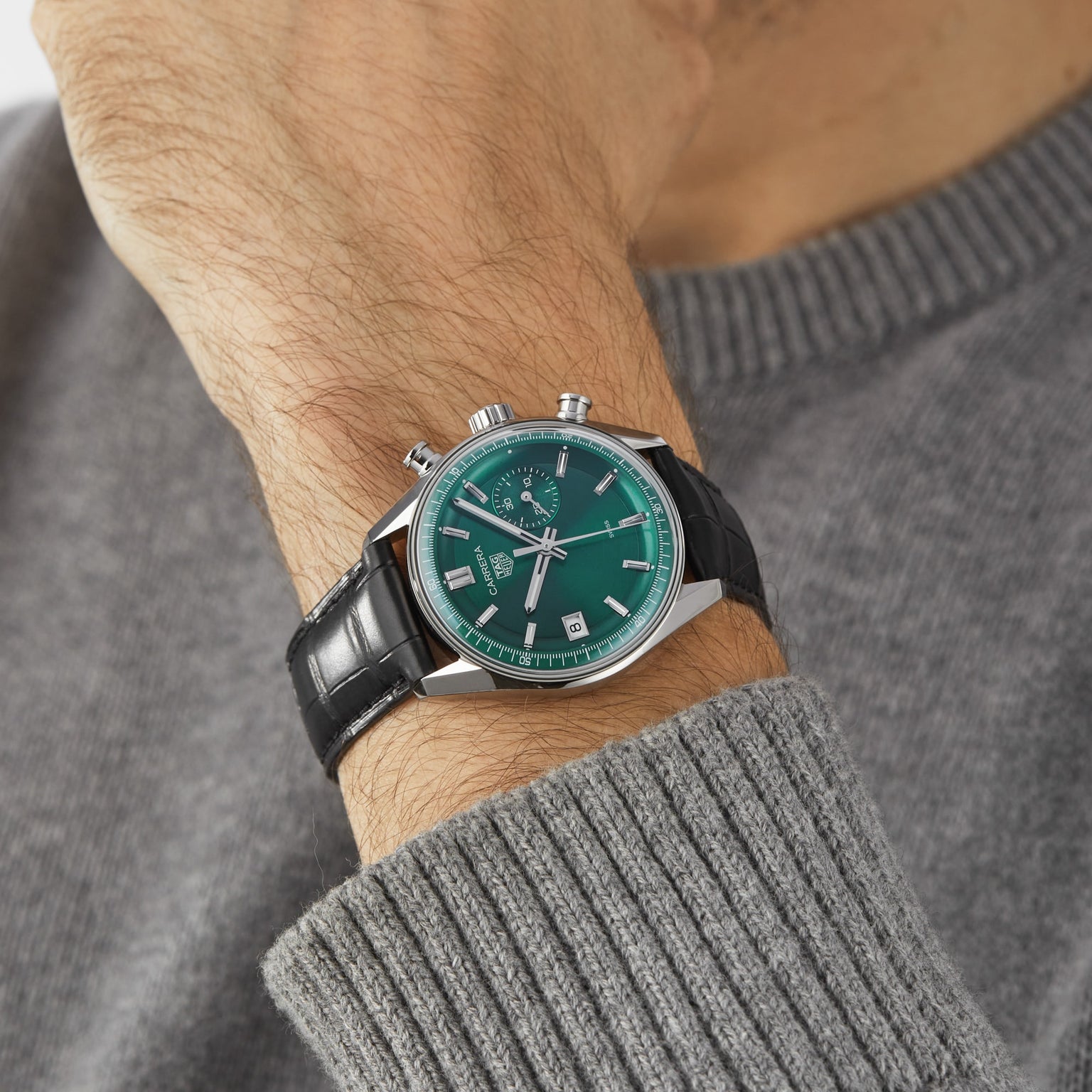 A person wearing a gray sweater and a TAG Heuer Carrera Chronograph wristwatch with a green face, black leather strap, and TH20-07 automatic movement.