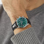 A person wearing a gray sweater and a TAG Heuer Carrera Chronograph wristwatch with a green face, black leather strap, and TH20-07 automatic movement.