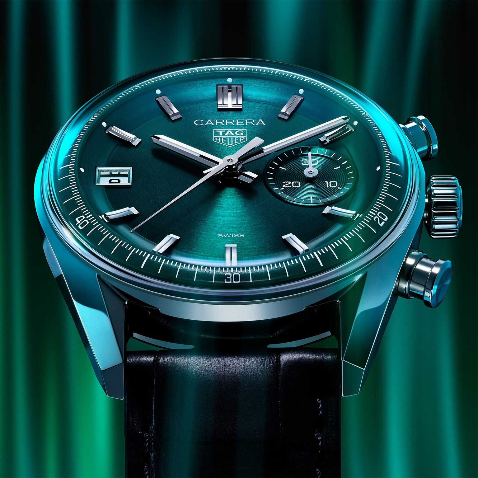 A close-up of the TAG Heuer Carrera Chronograph wristwatch, featuring a teal dial, date window, and chronograph on a black leather strap with metallic casing, set against a green background.