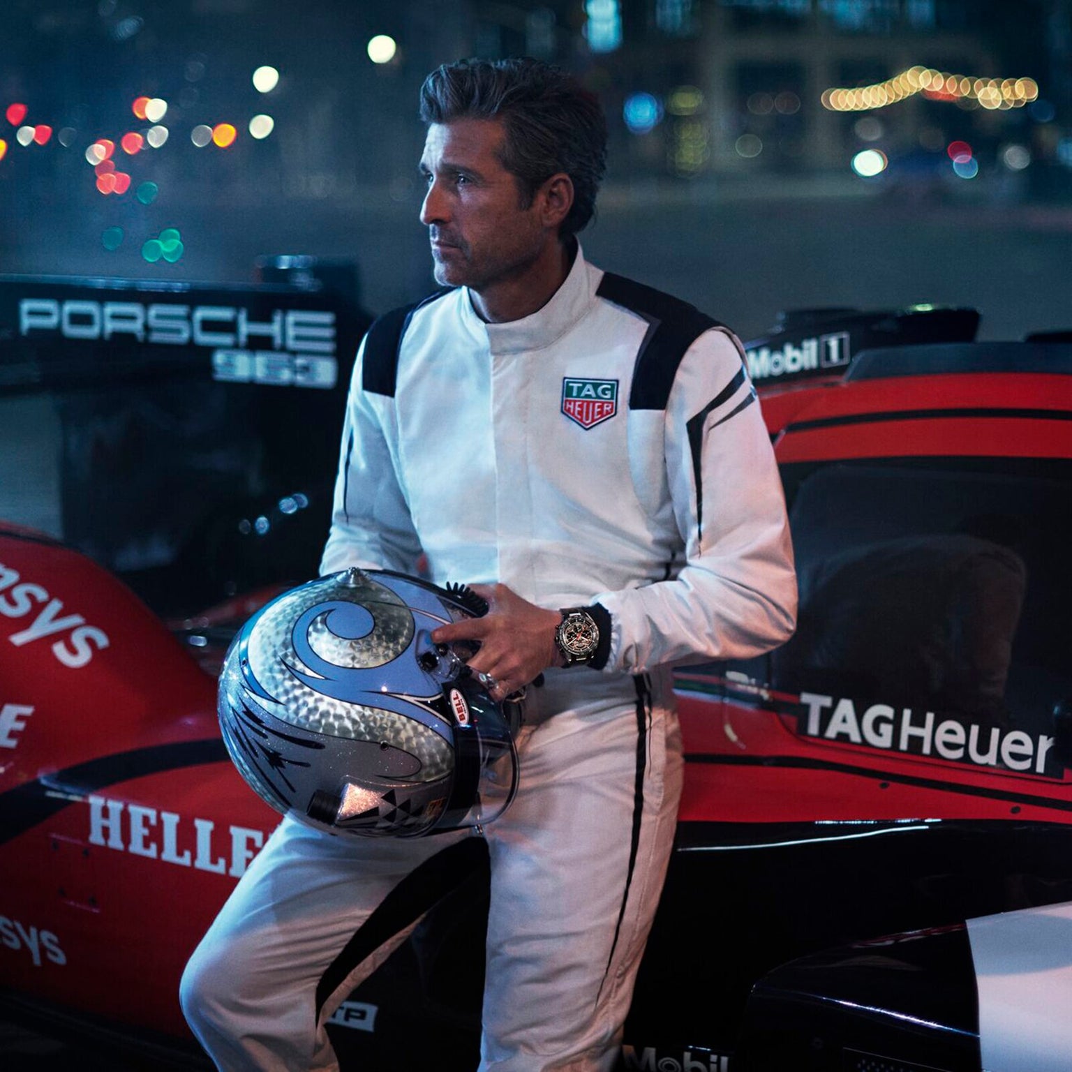 A man in a white racing suit stands by a Porsche race car at night, holding a helmet and wearing the TAG Heuer Limited Edition Carrera Chronograph x Porsche 963 watch.