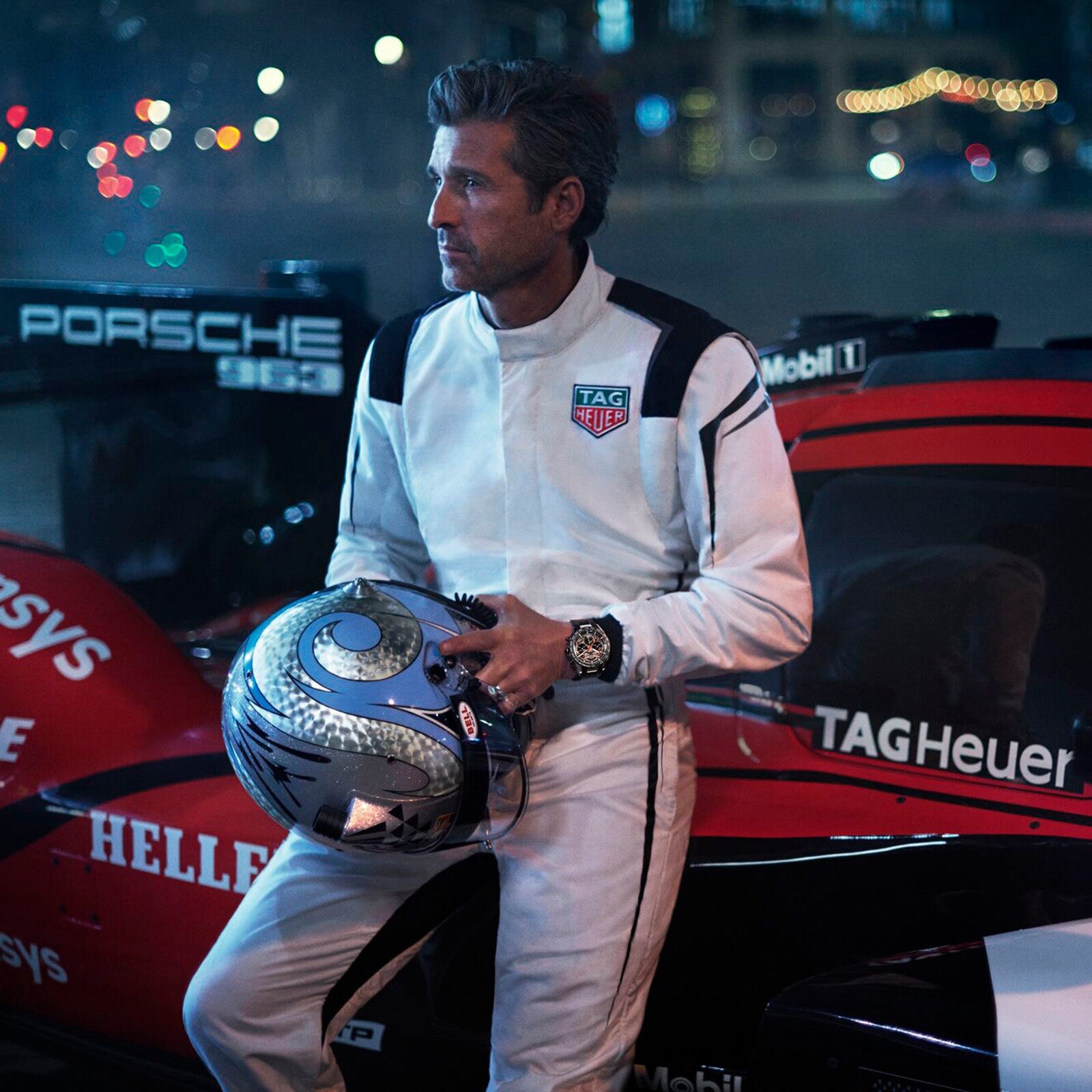 A man in a white racing suit stands by a Porsche race car at night, holding a helmet and wearing the TAG Heuer Limited Edition Carrera Chronograph x Porsche 963 watch.