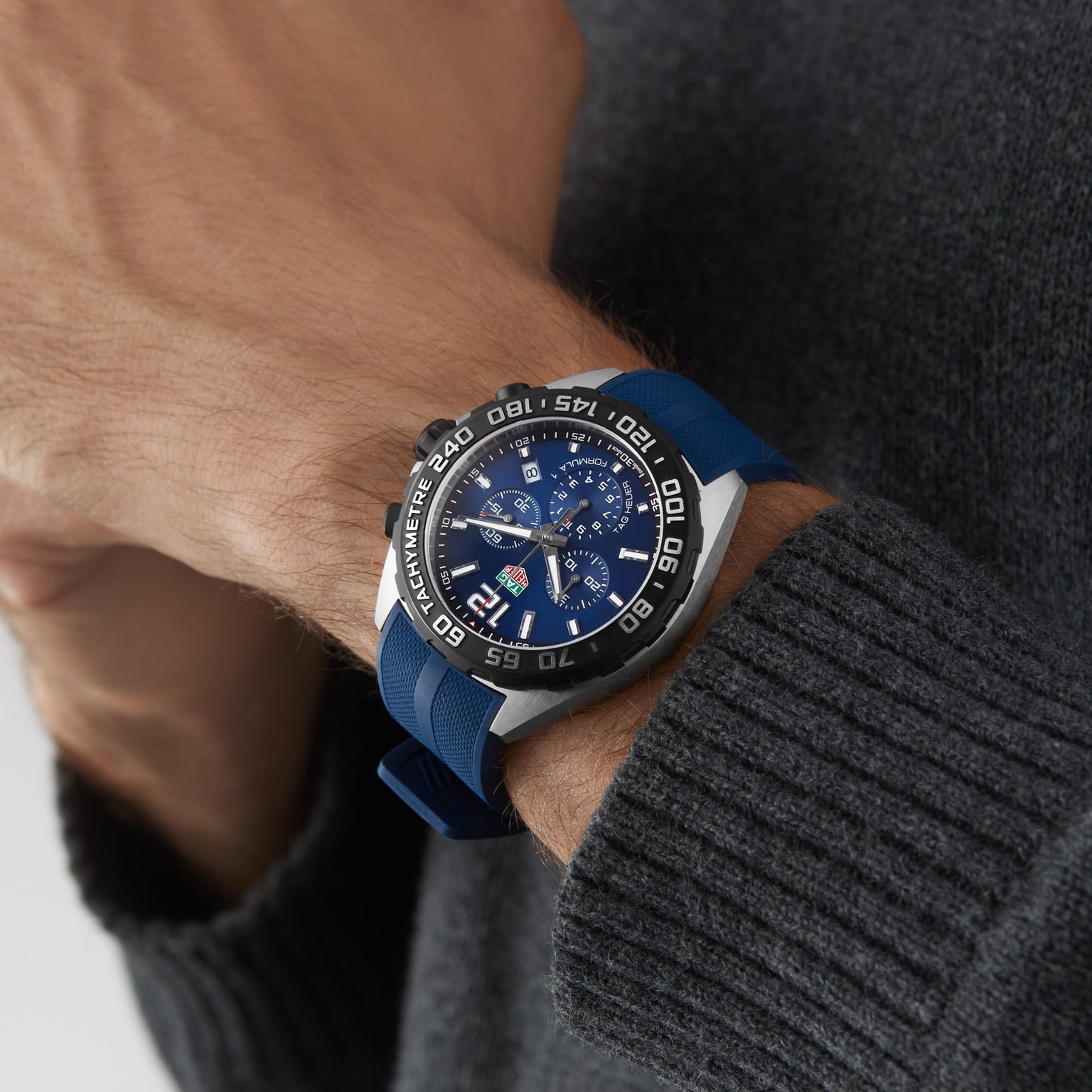 A person in a gray sweater showcases a TAG Heuer Formula 1 Chronograph with a blue and silver dial and blue strap on their left wrist.