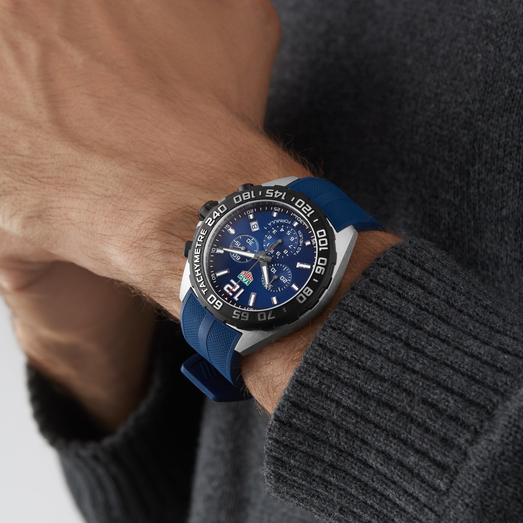 A person in a gray sweater showcases a TAG Heuer Formula 1 Chronograph with a blue and silver dial and blue strap on their left wrist.