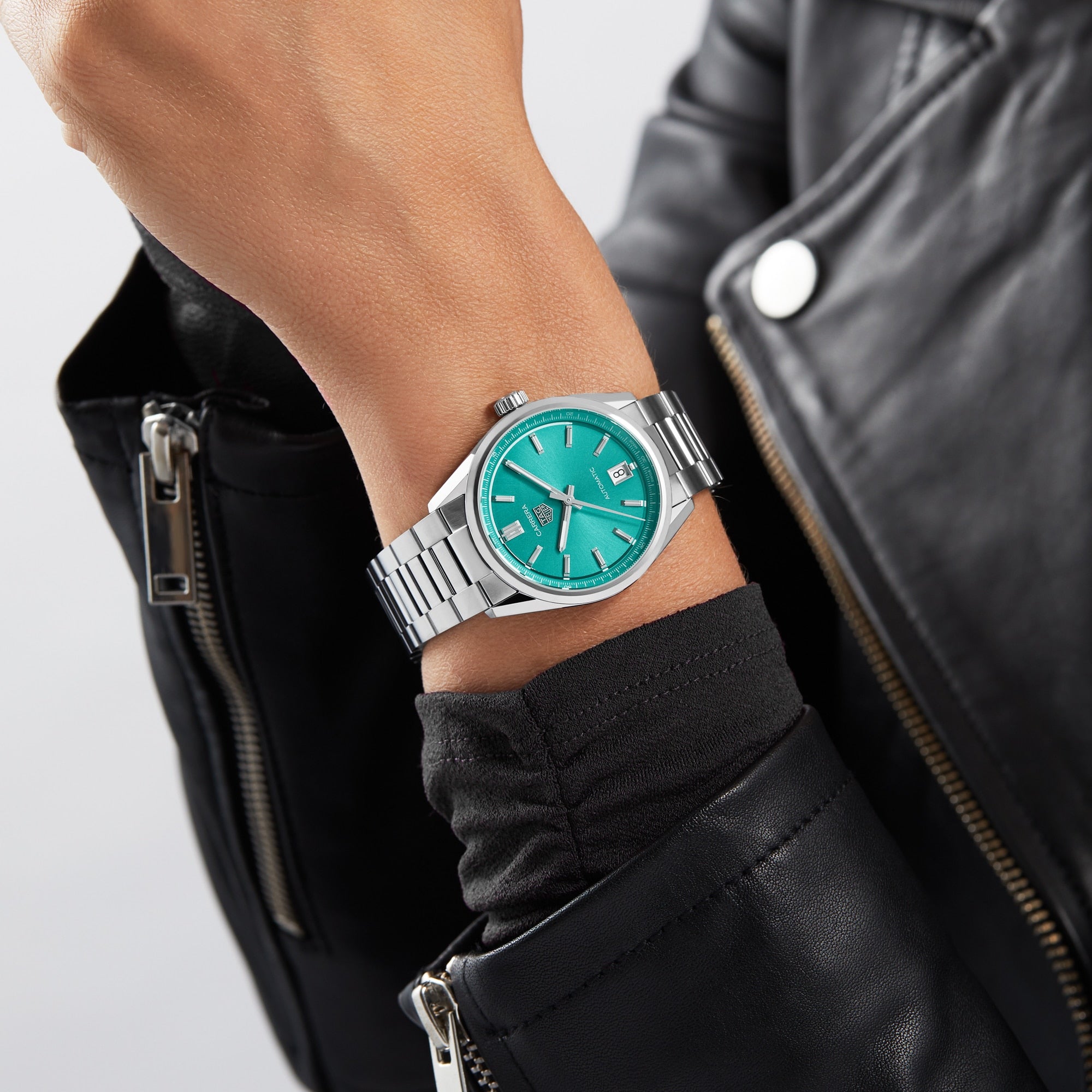 A person sporting a TAG Heuer Carrera Date—silver calibre 7 automatic watch with turquoise dial and date display—dons a black leather jacket, echoing Carrera style inspired by the legendary Panamericana race.