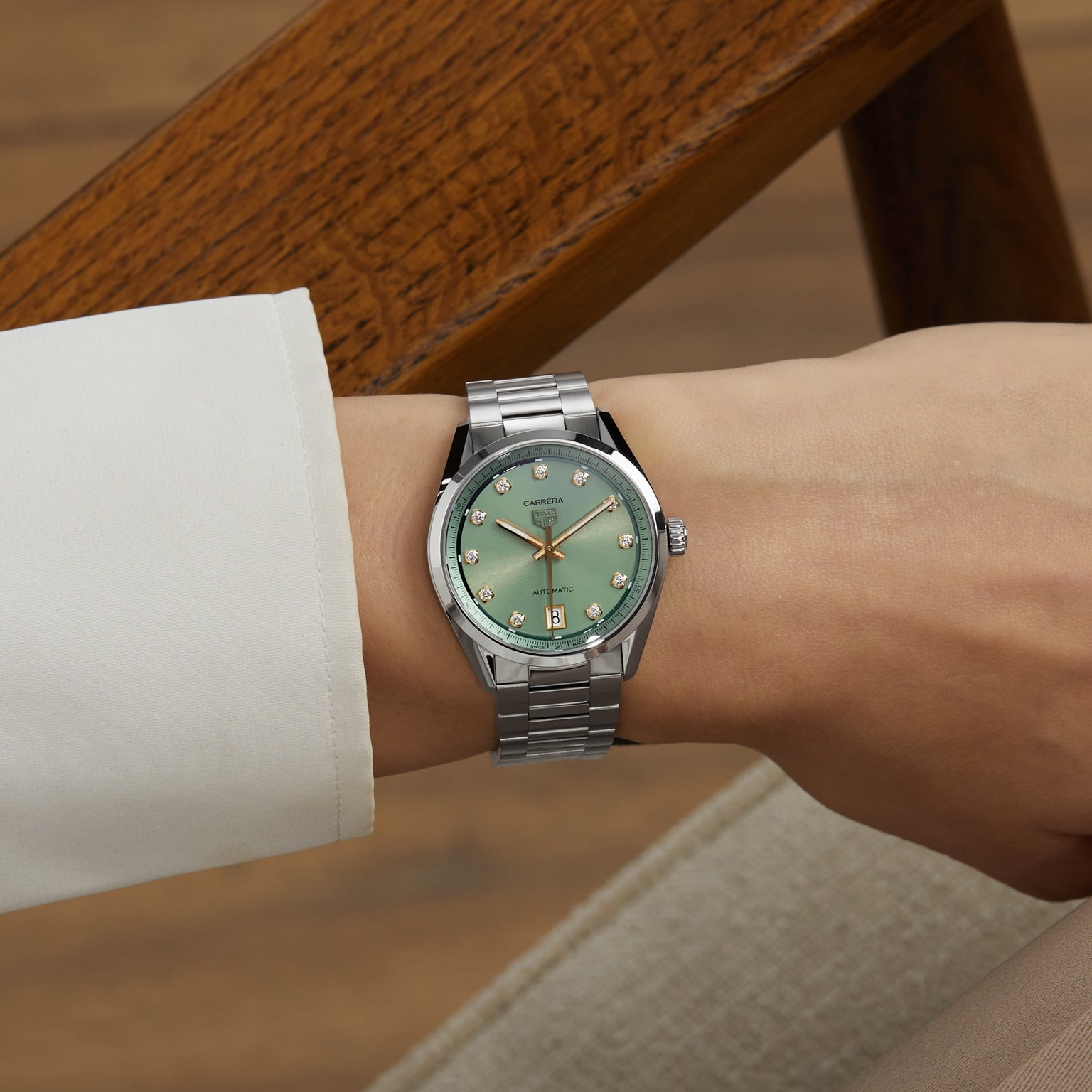 A person wearing a TAG Heuer Carrera Date watch with a green sunray-brushed dial and diamond hour markers sits on a beige chair with wooden armrests.