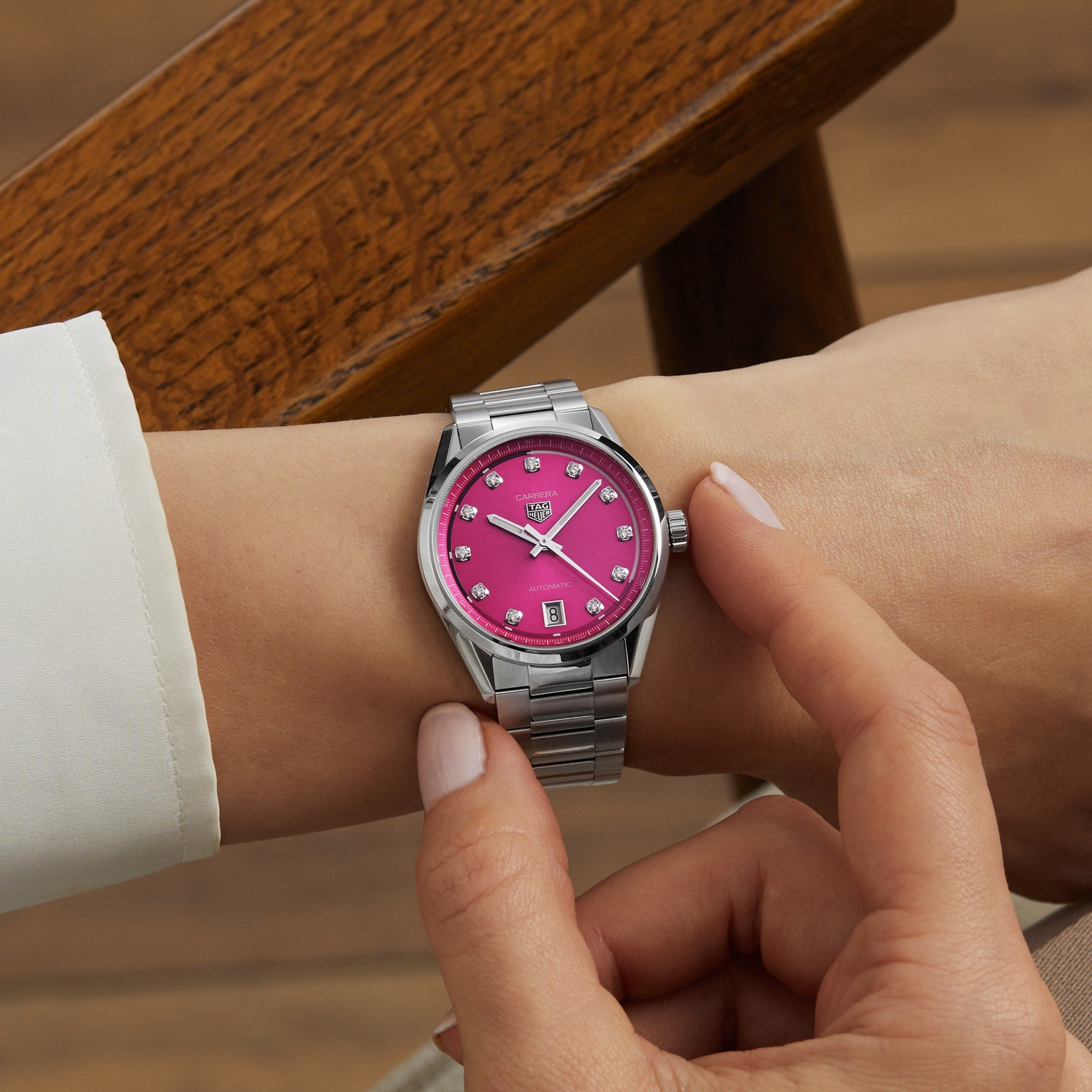 Someone adjusts a TAG Heuer Carrera Date watch, its pink snail-brushed dial and VS diamonds gleaming as their arm rests on a wooden chair.