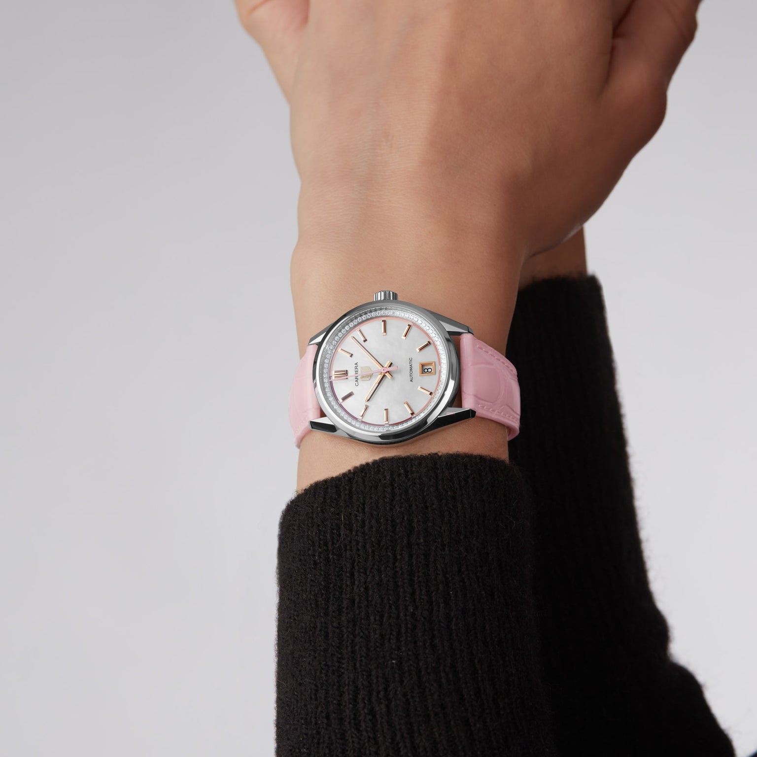 A person wears a TAG Heuer Carrera Calibre 7 silver wristwatch with a mother-of-pearl dial and rose gold-plated indexes on their left wrist against a plain background.