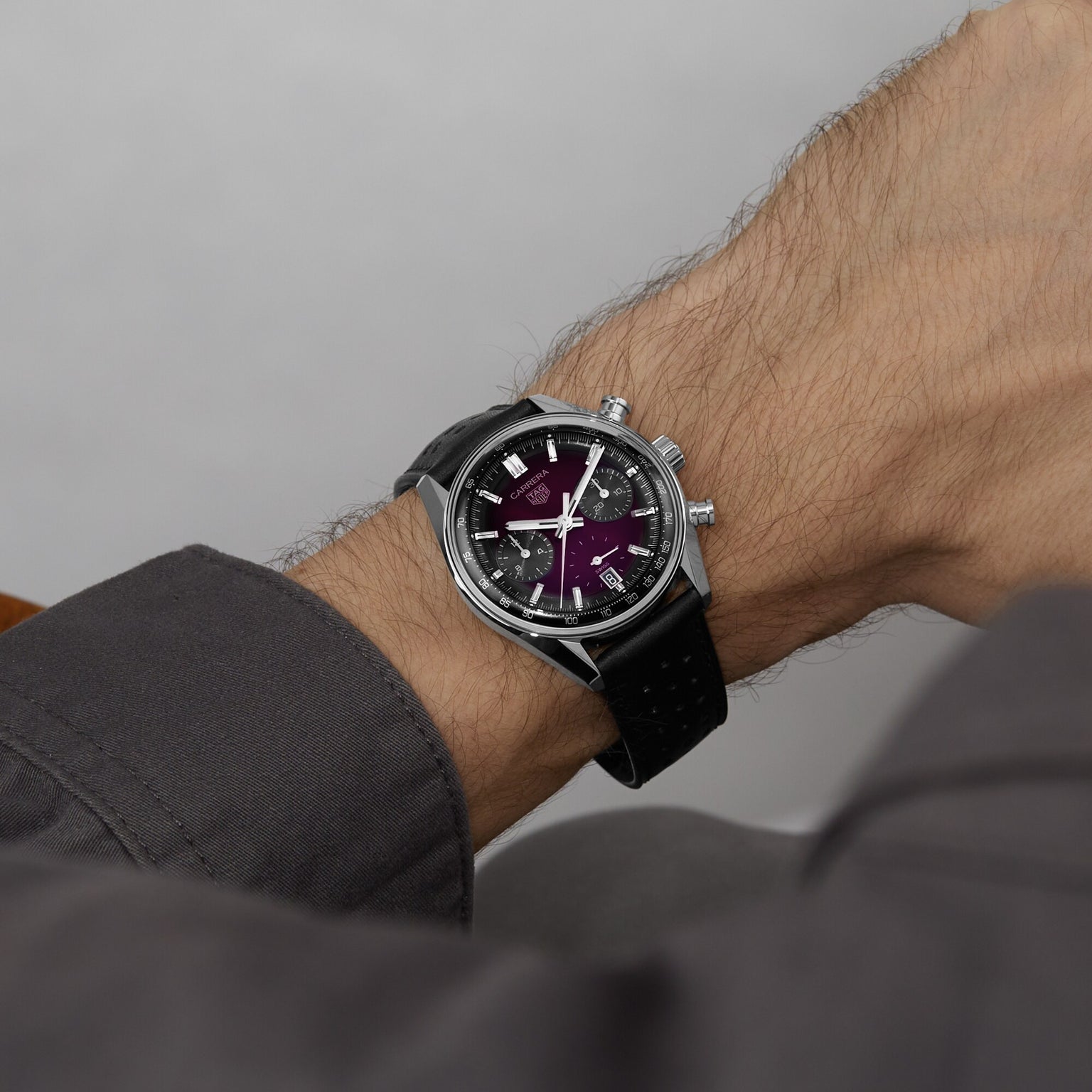 A close-up shows a person’s wrist wearing the TAG Heuer Carrera Chronograph, featuring a silver case, black strap, and a smoky purple sunray brushed dial.