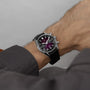 A close-up shows a person’s wrist wearing the TAG Heuer Carrera Chronograph, featuring a silver case, black strap, and a smoky purple sunray brushed dial.