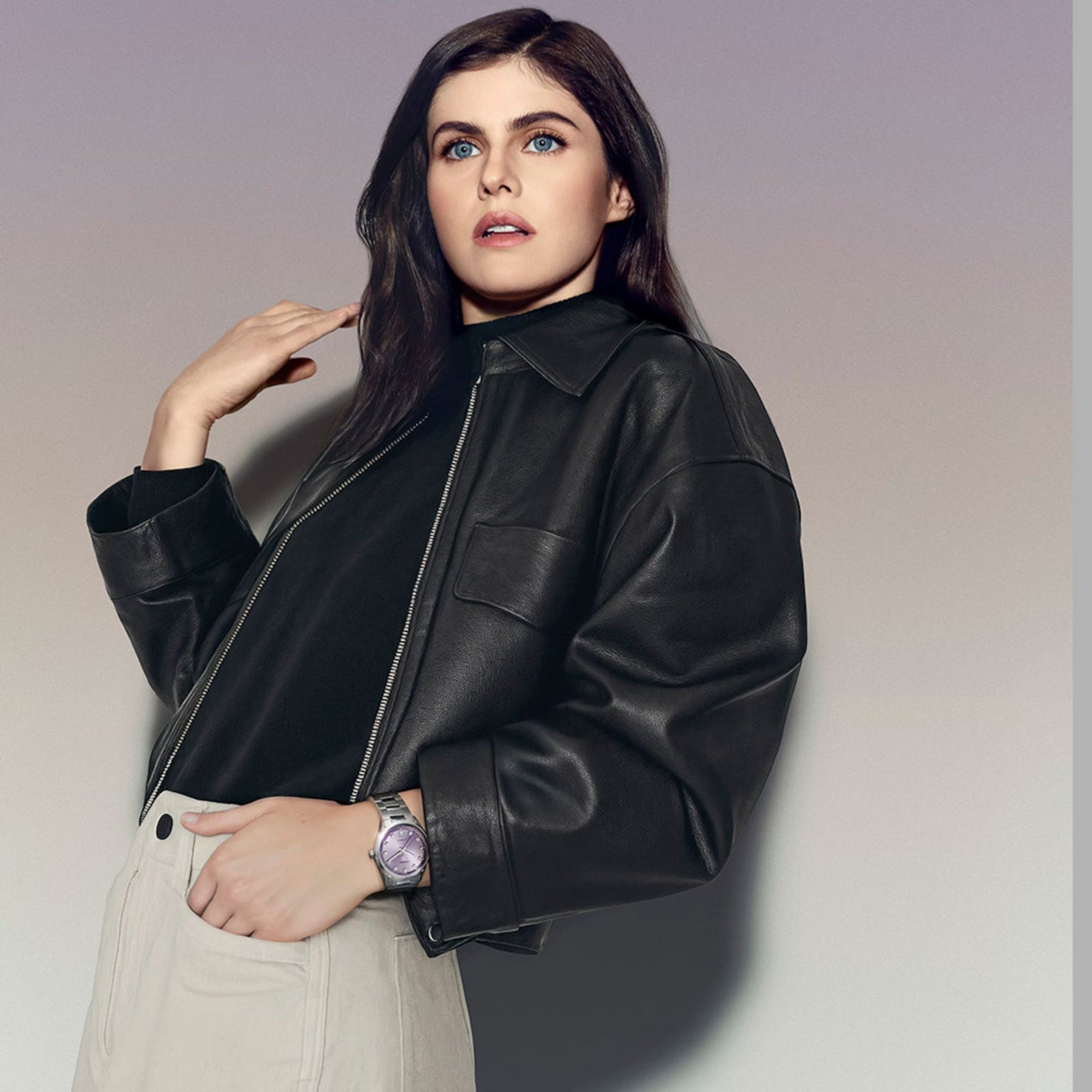 Woman with long dark hair in a black leather jacket and beige pants, glancing upward, showcases the TAG Heuer Carrera Date watch.