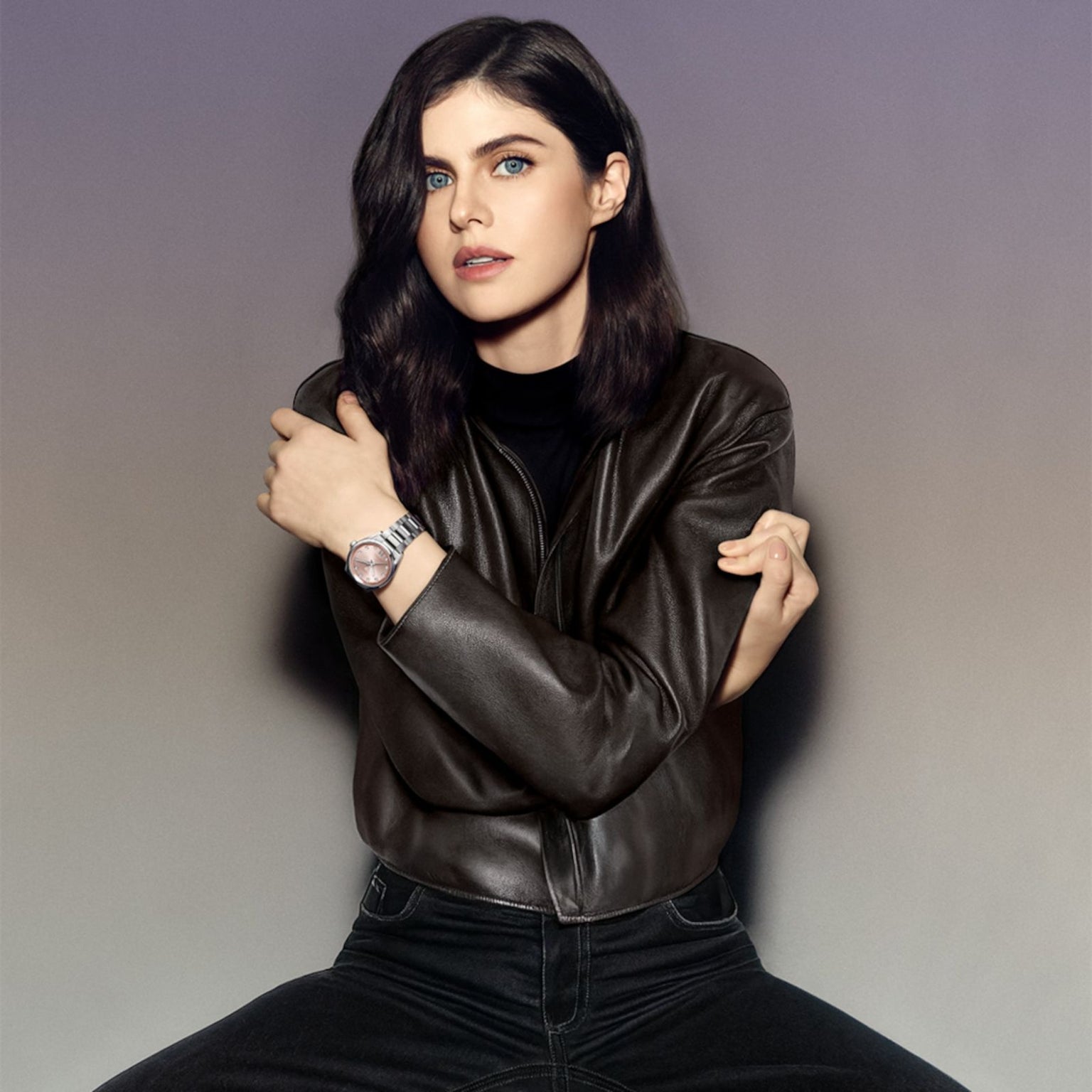 Woman with long dark hair in a leather jacket, seated with arms crossed, wearing the TAG Heuer Carrera Date watch.