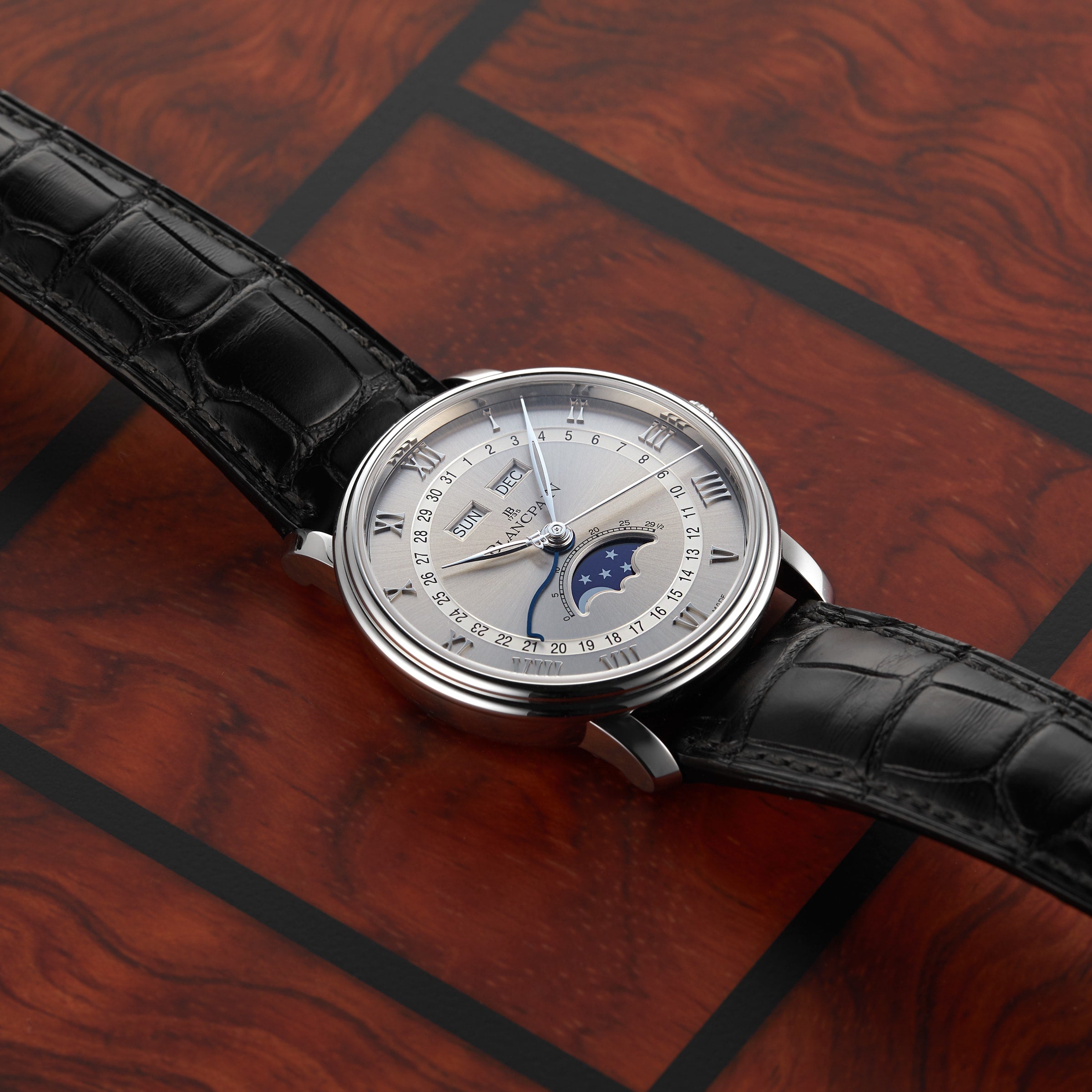 The Blancpain Villeret Quantième Complet luxury wristwatch, featuring a black leather strap and moonphase dial, is displayed on a wooden surface.