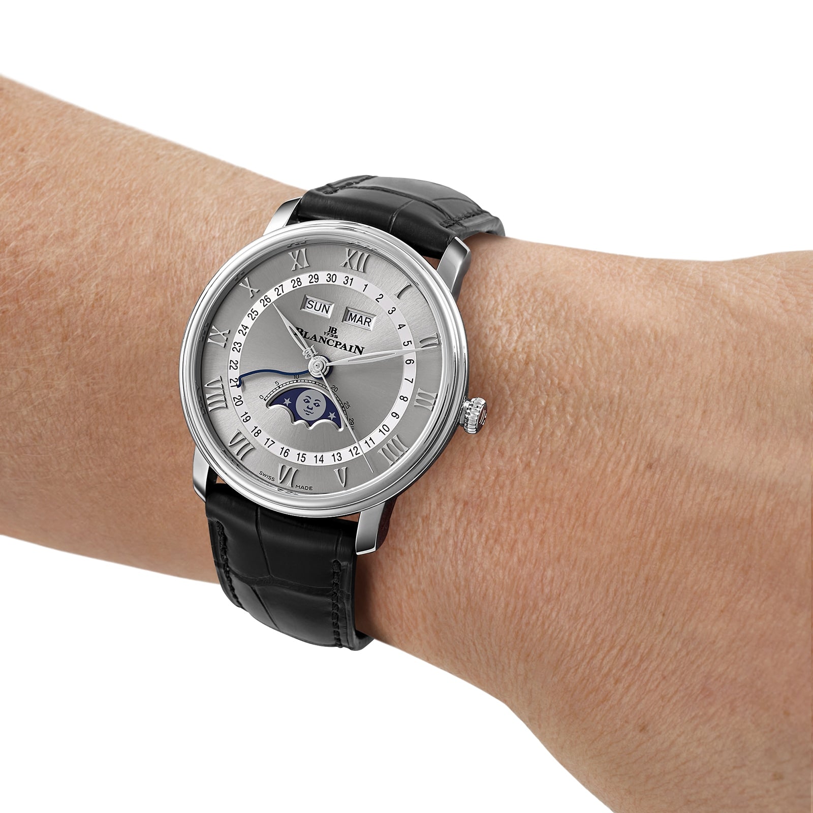 A Blancpain Villeret Quantième Complet silver wristwatch with a black leather band, moonphase dial, and Roman numerals on a persons wrist.