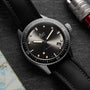 A close-up of a Blancpain Fifty Fathoms Bathyscaphe diver’s watch with a black dial, black strap, and date window, featuring an automatic movement, placed on a surface beside part of a map and a metal tool.