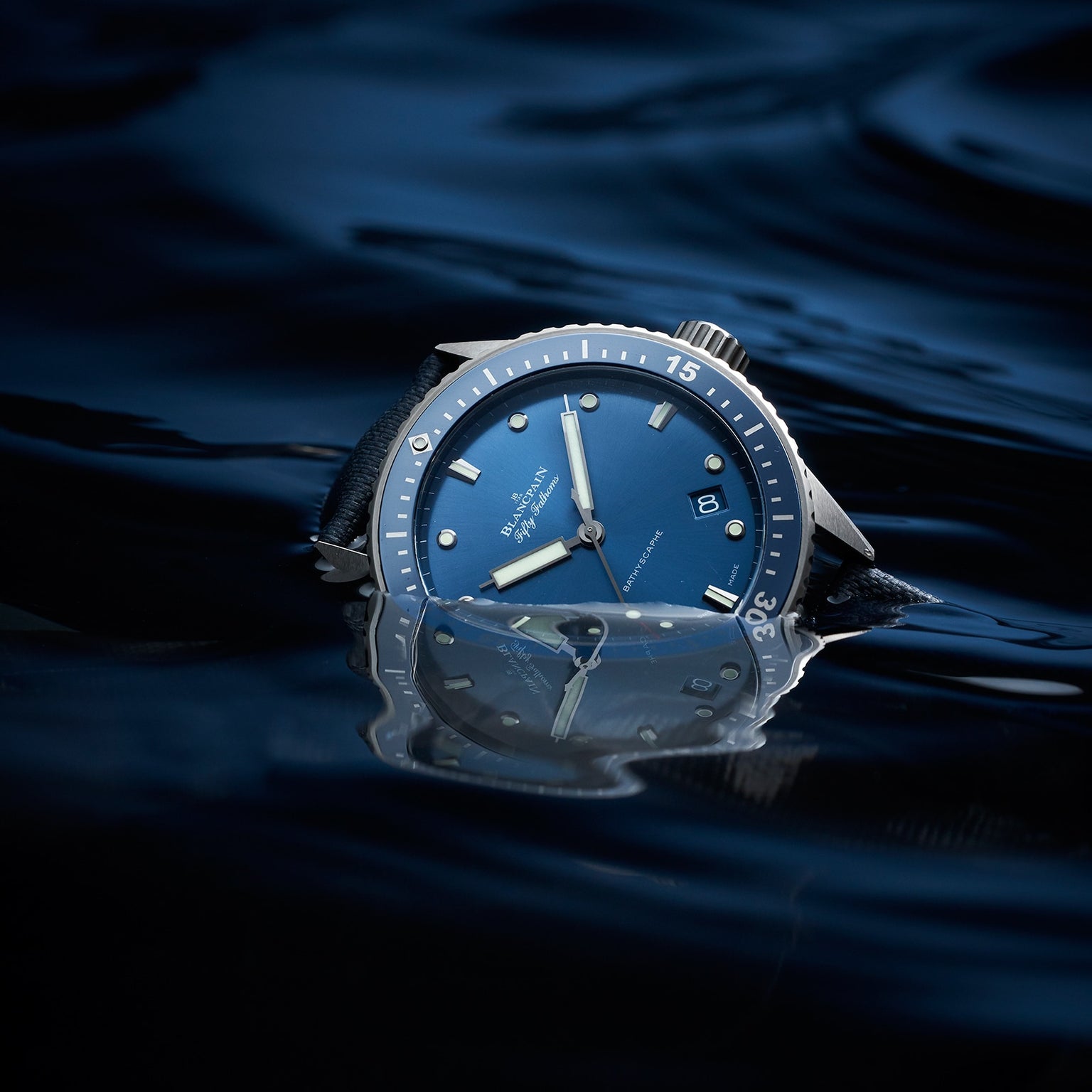 The Blancpain Fifty Fathoms Bathyscaphe, an automatic divers watch with a blue dial and rotating bezel, is partially submerged in dark water, its reflection shimmering on the surface.