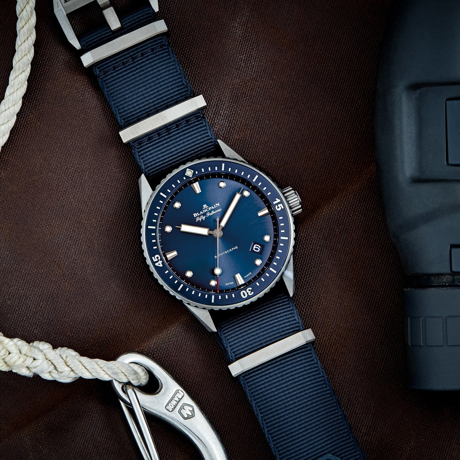 The Blancpain Fifty Fathoms Bathyscaphe, featuring a blue face and blue strap, rests on a brown surface beside a coiled white rope, a metal carabiner, and a black object.