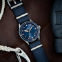 The Blancpain Fifty Fathoms Bathyscaphe, featuring a blue face and blue strap, rests on a brown surface beside a coiled white rope, a metal carabiner, and a black object.
