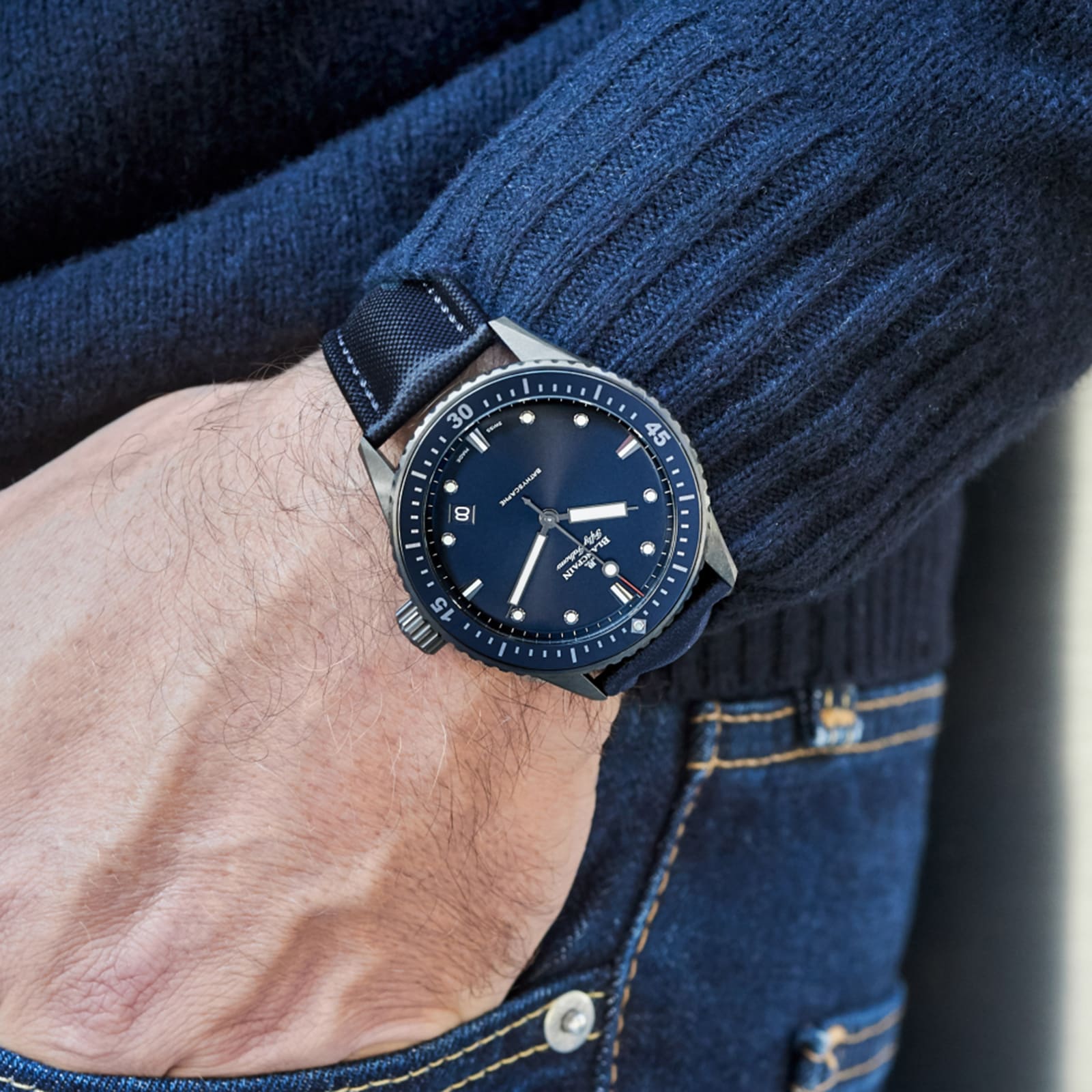 A person in a dark blue sweater and jeans wears a Blancpain Fifty Fathoms Bathyscaphe black divers watch with a fabric strap and automatic movement on their left wrist.