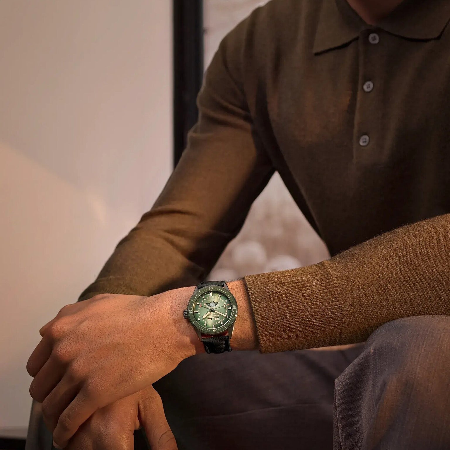 A person in a brown long-sleeve polo and gray pants is seated, displaying a Blancpain Fifty Fathoms Bathyscaphe Quantième Complet Phases de Lune green analog wristwatch on their left wrist.