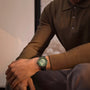 A person in a brown long-sleeve polo and gray pants is seated, displaying a Blancpain Fifty Fathoms Bathyscaphe Quantième Complet Phases de Lune green analog wristwatch on their left wrist.