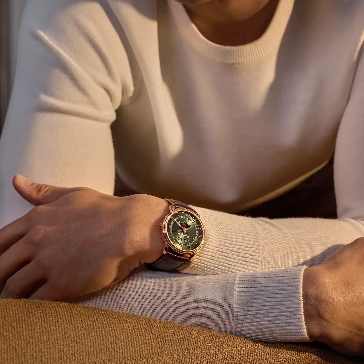 A person in a cream sweater rests their arms on a brown surface, showcasing the Blancpain Villeret Quantième Complet wristwatch with a green dial and leather strap.