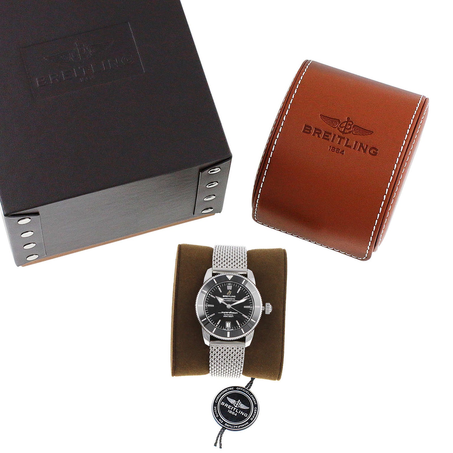 The Breitling Superocean Heritage B20 Automatic 42 is shown with a mesh stainless steel bracelet on a cushion, alongside a black box, brown leather case, and tag.