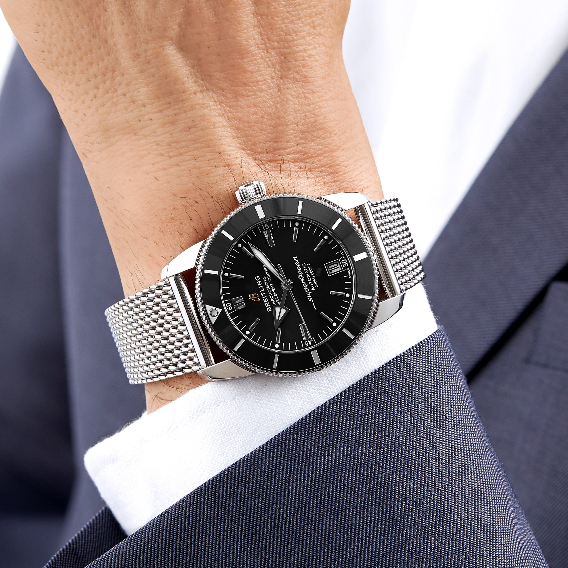 A person in a navy blue suit jacket and white shirt wears a Breitling Superocean Heritage B20 Automatic 42 wristwatch with a stainless steel bracelet, black dial, and bezel.
