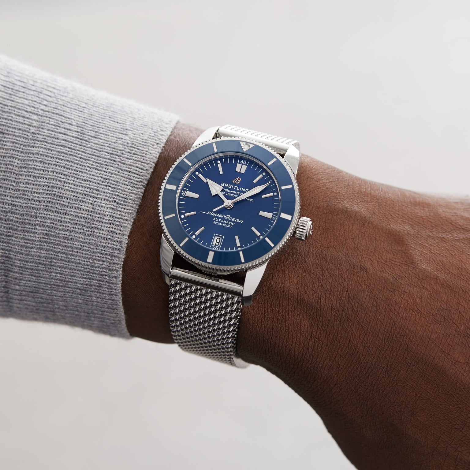 A person wears a Breitling Superocean Heritage B20 Automatic 42 with a silver mesh strap, gun blue ceramic bezel, stainless steel case, blue dial displaying 10:09, and a date window at 6 o’clock.