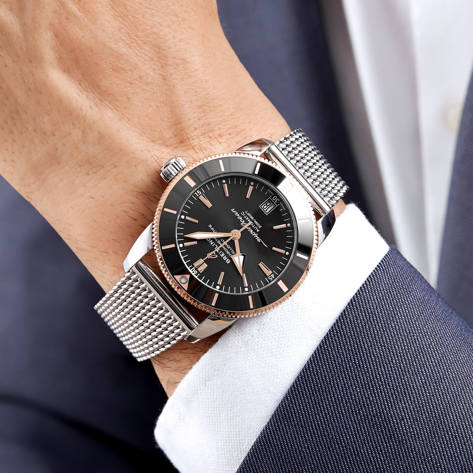 A person wearing a Breitling Superocean Heritage B20 Automatic 42 with a black dial and mesh strap on their left wrist, dressed in a dark blue suit jacket and white shirt.