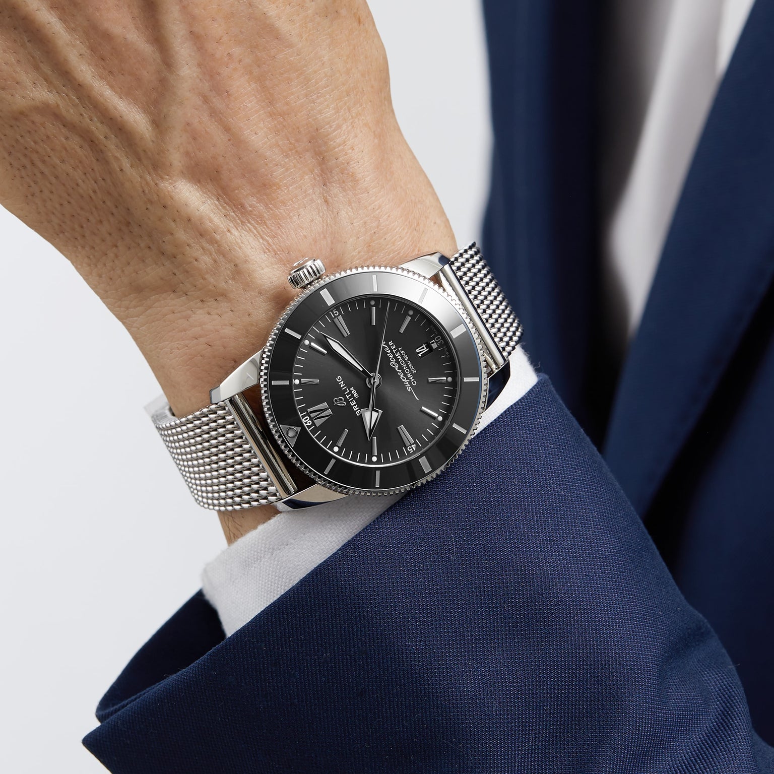A person in a navy blue suit wears a Breitling Superocean Heritage B20 Automatic 44 wristwatch with a black dial, date display, and stainless steel bracelet.