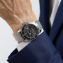 A person in a navy blue suit wears a Breitling Superocean Heritage B20 Automatic 44 wristwatch with a black dial, date display, and stainless steel bracelet.