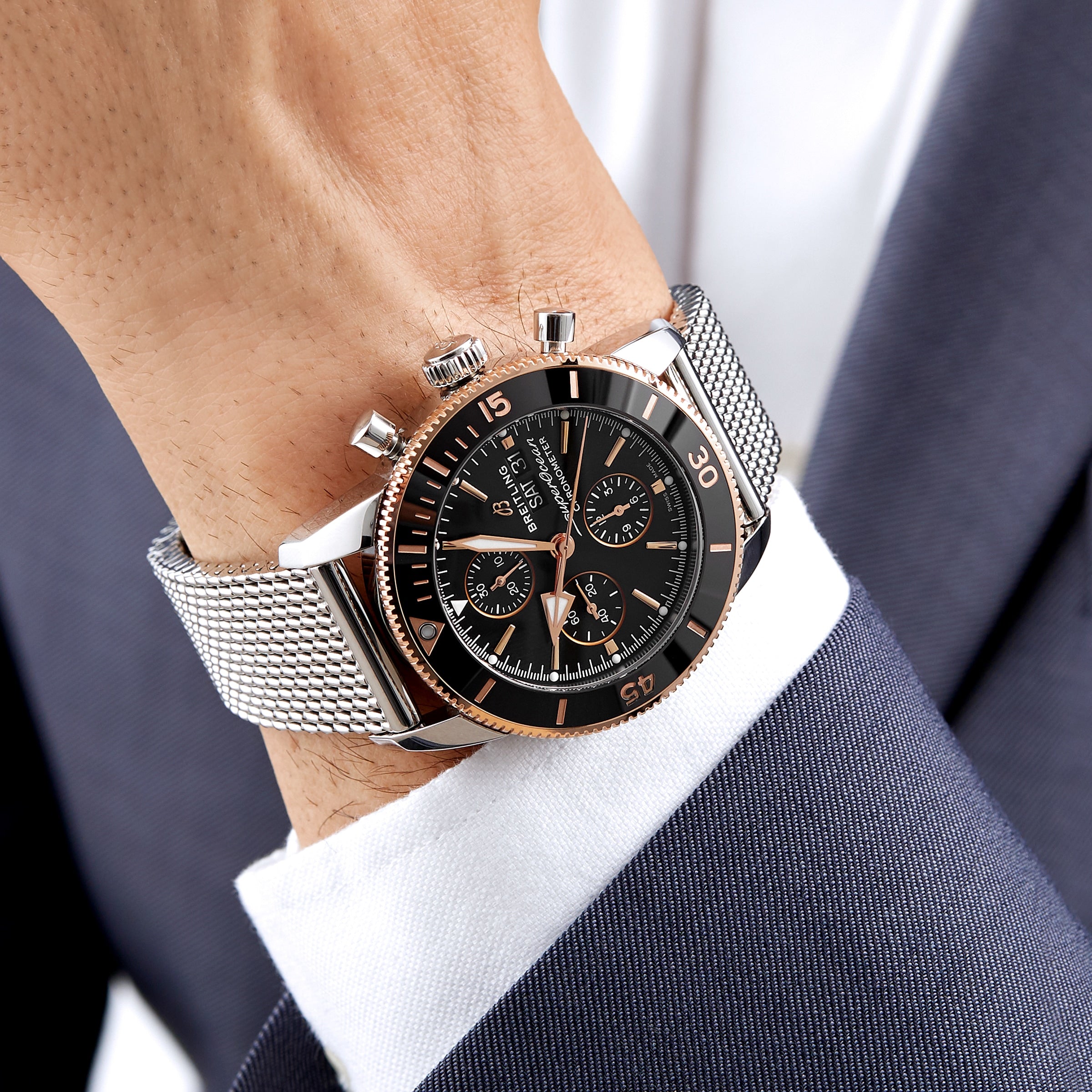 A close-up of a person in a navy suit and white shirt wearing a Breitling Superocean Heritage B20 Automatic 44 with a stainless steel case, black dial, and mesh strap.