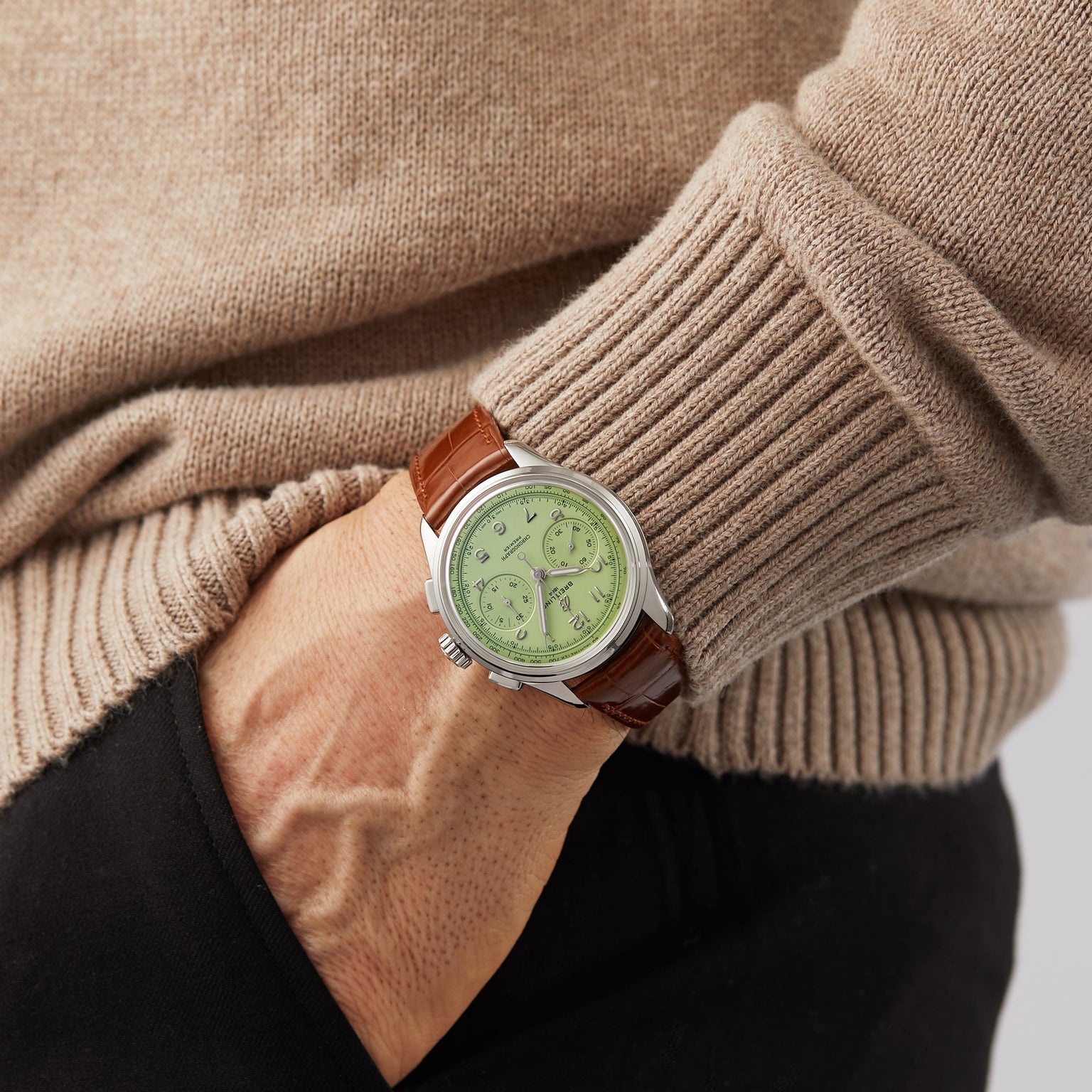 A person wearing a beige sweater and black pants sports the Breitling Premier B09 Chronograph 40 with a brown leather strap, green dial, chronograph features, and precise B09 movement—hand in pocket.