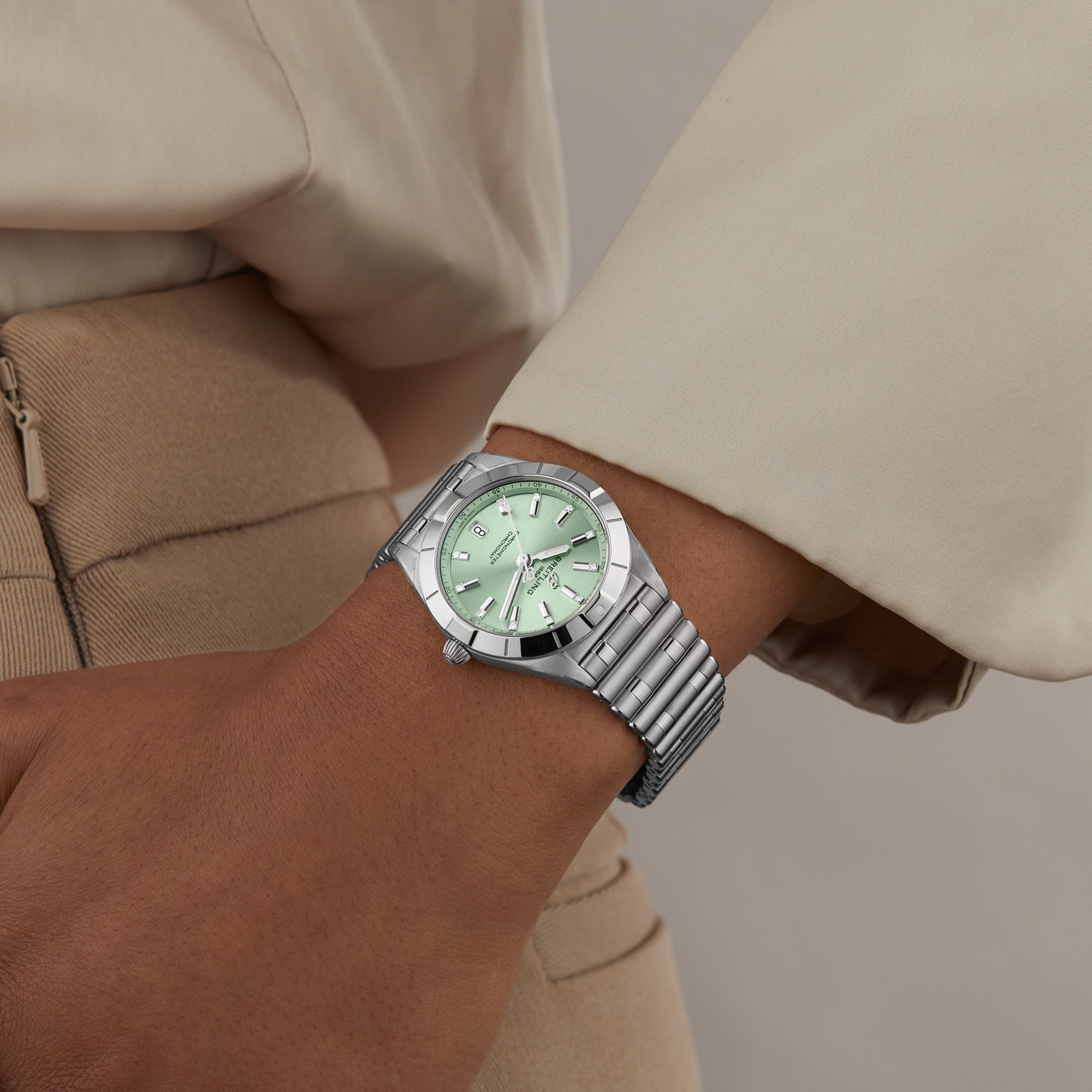 A person in beige attire wears a Breitling Chronomat 32, featuring a silver case, light green dial, and metal Rouleaux bracelet.