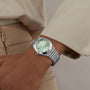 A person in beige attire wears a Breitling Chronomat 32, featuring a silver case, light green dial, and metal Rouleaux bracelet.