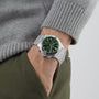 Wearing a Breitling Chronomat B01 42 with a green dial and stainless steel case, paired with a grey sweater and green pants, a person stands casually with one hand in their pocket.