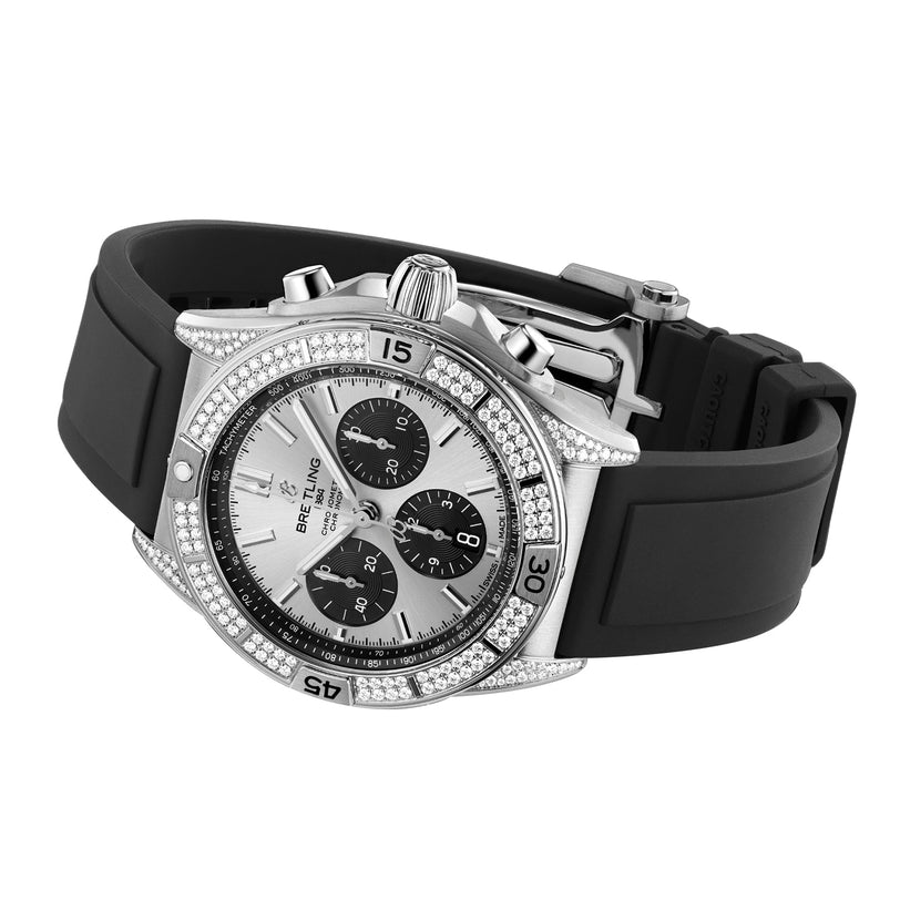 The Breitling Chronomat B01 42 is a silver chronograph watch with a stainless steel case, diamond-encrusted bezel, black subdials, and a black rubber strap.