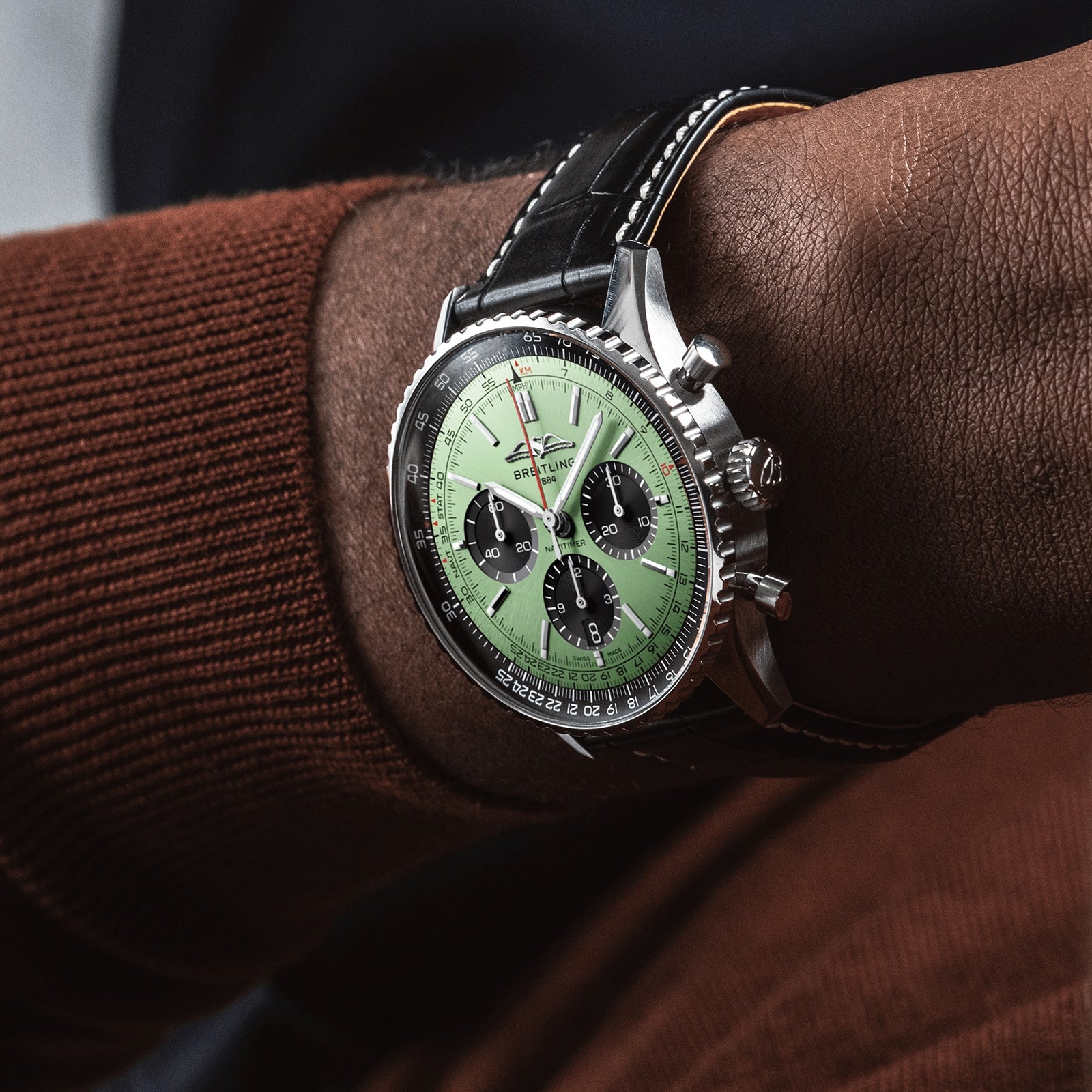 A close-up of a person wearing a Breitling Navitimer B01 Chronograph 43 with a green dial, three subdials, and black leather strap on a brown-sleeved arm.