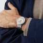 A person in a beige sweater and navy blue jacket with one arm folded across the chest wears a Breitling Navitimer B01 Chronograph 41 with a silver case and white dial.