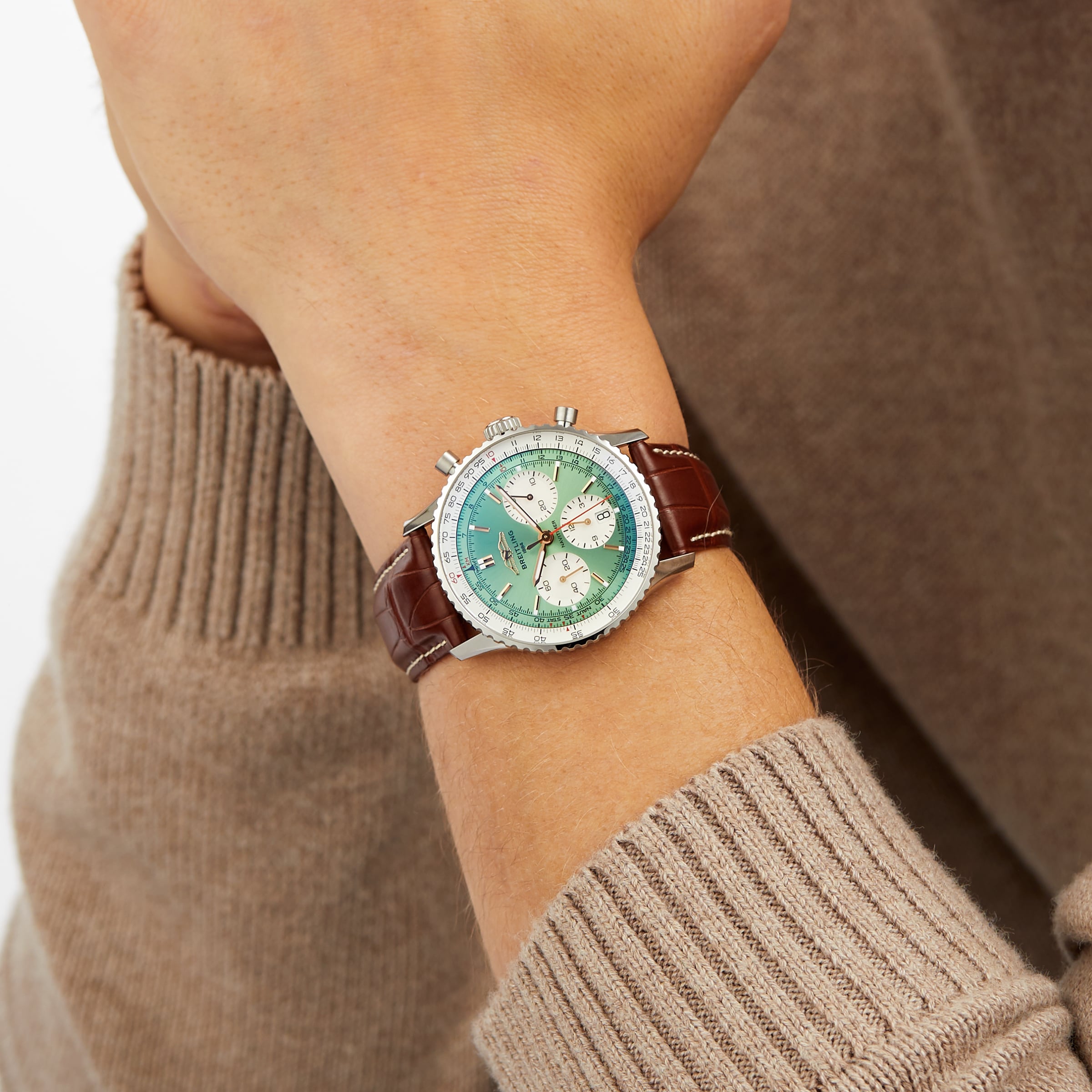 A person in a brown long-sleeve sweater showcases the Breitling Navitimer B01 Chronograph 41, featuring a green dial, white subdials, and a brown leather strap.