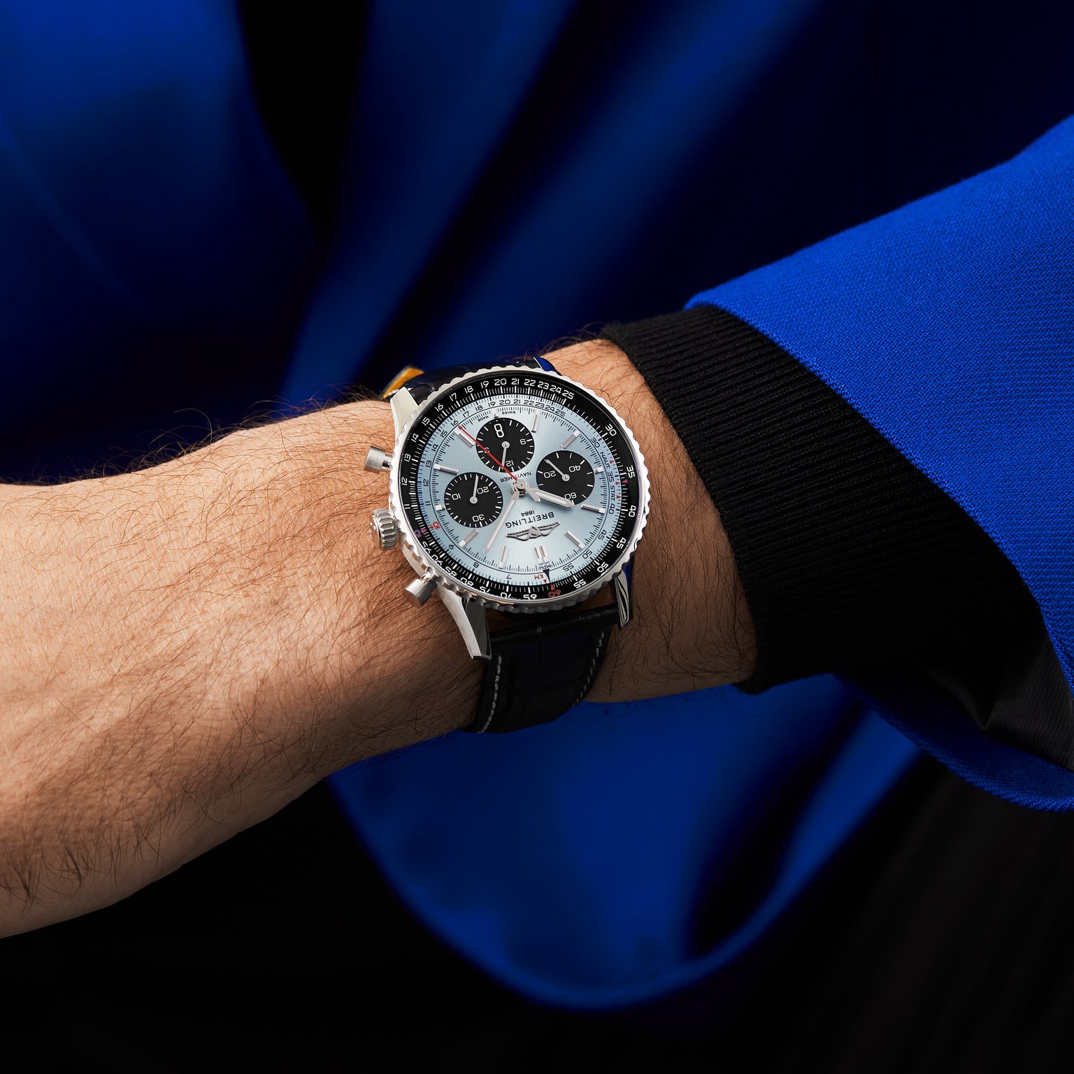 Wearing a blue and black jacket, a person shows off a Breitling Navitimer B01 Chronograph 43 with a white and black dial and black leather strap on their left wrist.