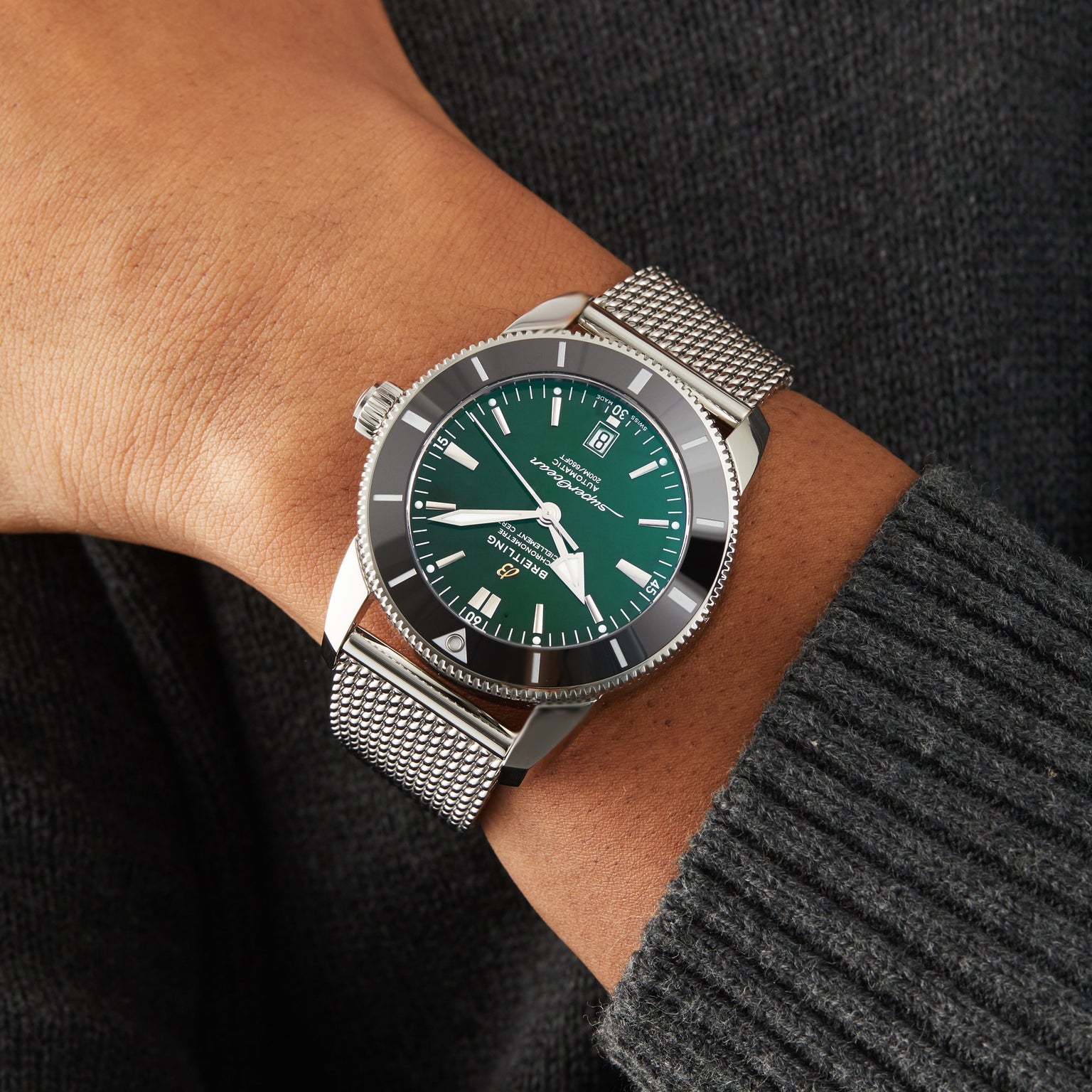 A person wears a Breitling Superocean Heritage B20 Automatic 46, featuring a green dial, silver mesh band, and 46mm stainless steel case, over a dark ribbed sweater.