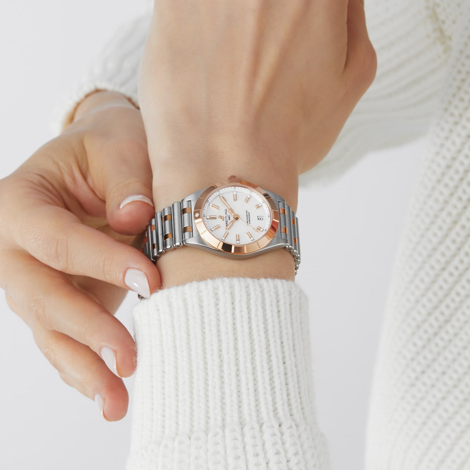 A person adjusts their Breitling Chronomat 32 wristwatch in silver and gold with a white dial and metal band over the sleeve of a white knit sweater.