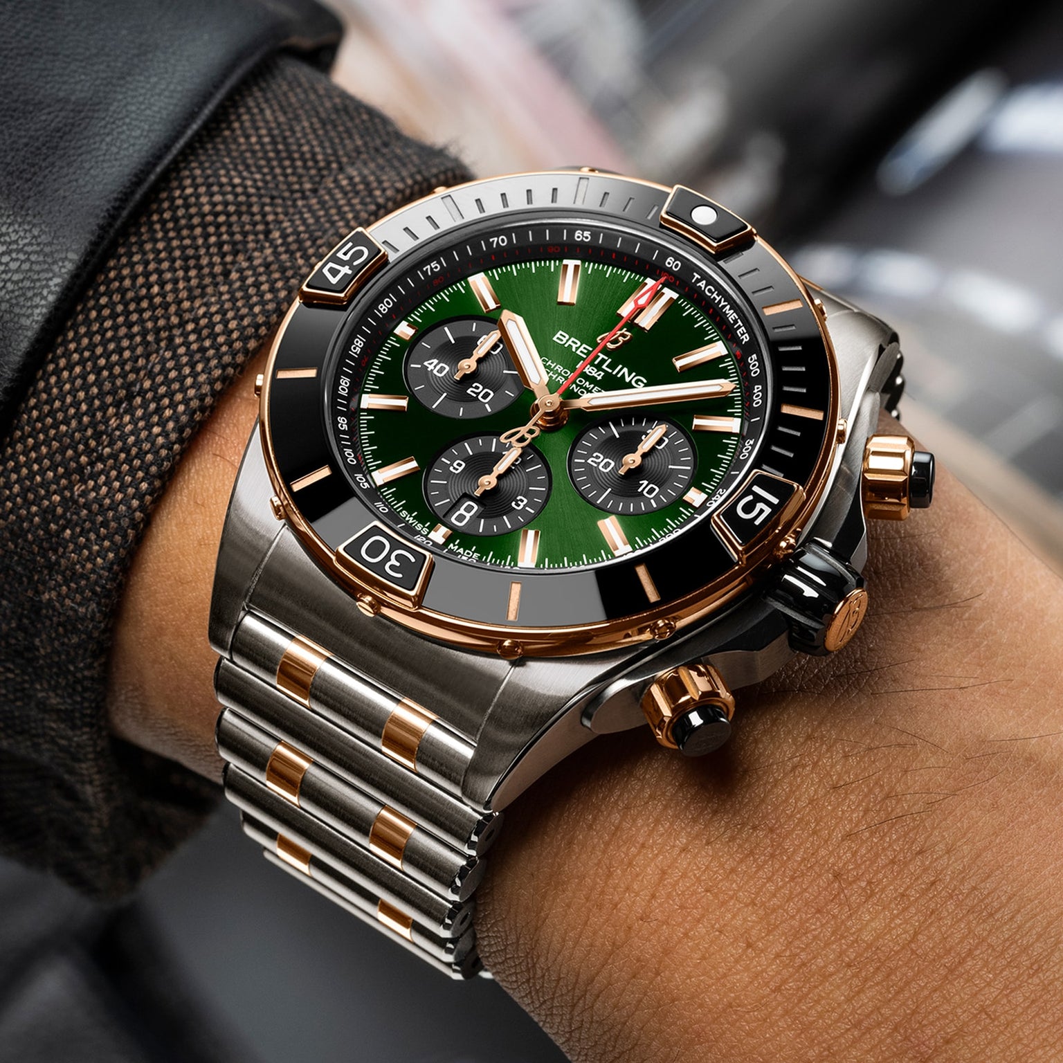 A close-up of a Breitling Super Chronomat B01 44 chronograph wristwatch with an emerald dial, gold accents, and metal bracelet on a person’s wrist, powered by the Breitling Manufacture 01 movement.